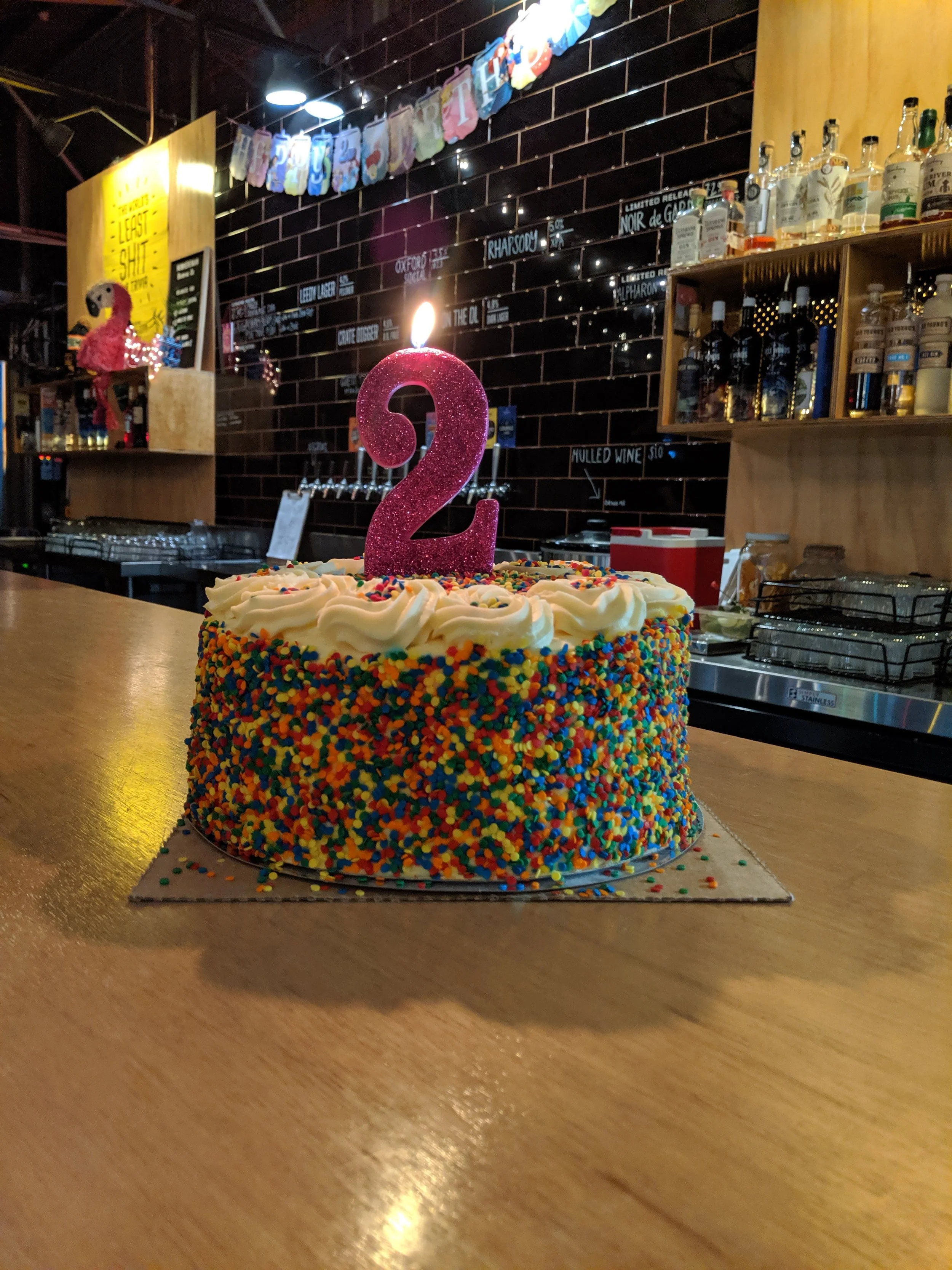 A birthday cake with rainbow sprinkles and white frosting, topped with a pink glittery number two candle, on a wooden table in a bar or cafe setting.
