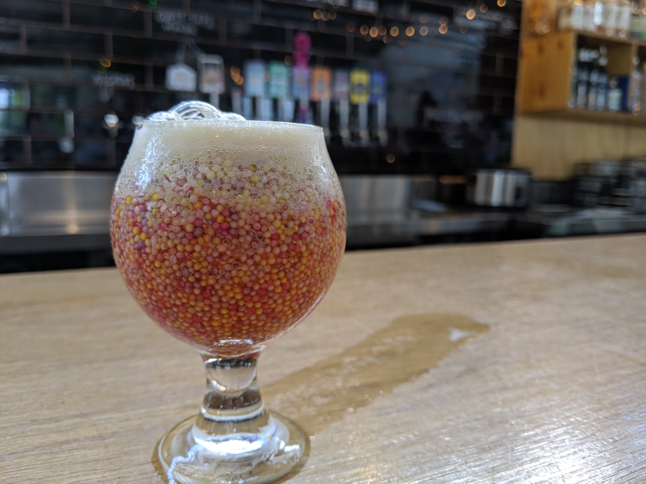 A glass of beer with a foamy head, filled with multicolored tiny round beads, sitting on a wooden bar counter in a bar or restaurant.