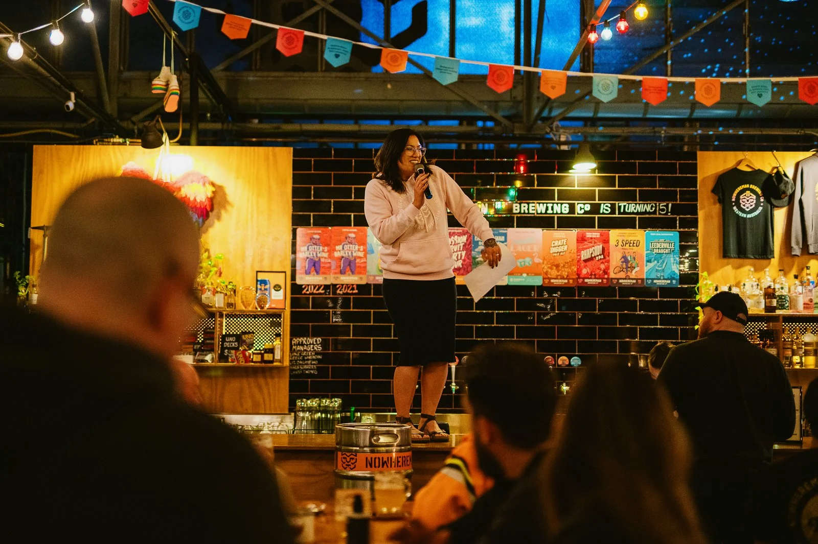 A woman standing on a stage with a microphone, speaking to an audience in a bar or brewery. The background features a brick wall with posters, colorful banners hanging from the ceiling, and shelves with bottles. The scene is warmly lit and the audien