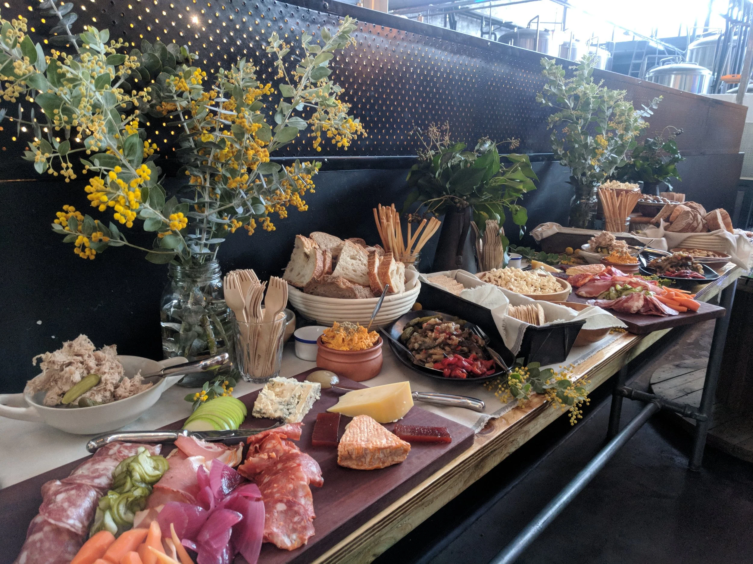 A buffet table with assorted foods including bread, cheese, meats, salads, and vegetables, decorated with green leafy plants and yellow flowers in glass jars.