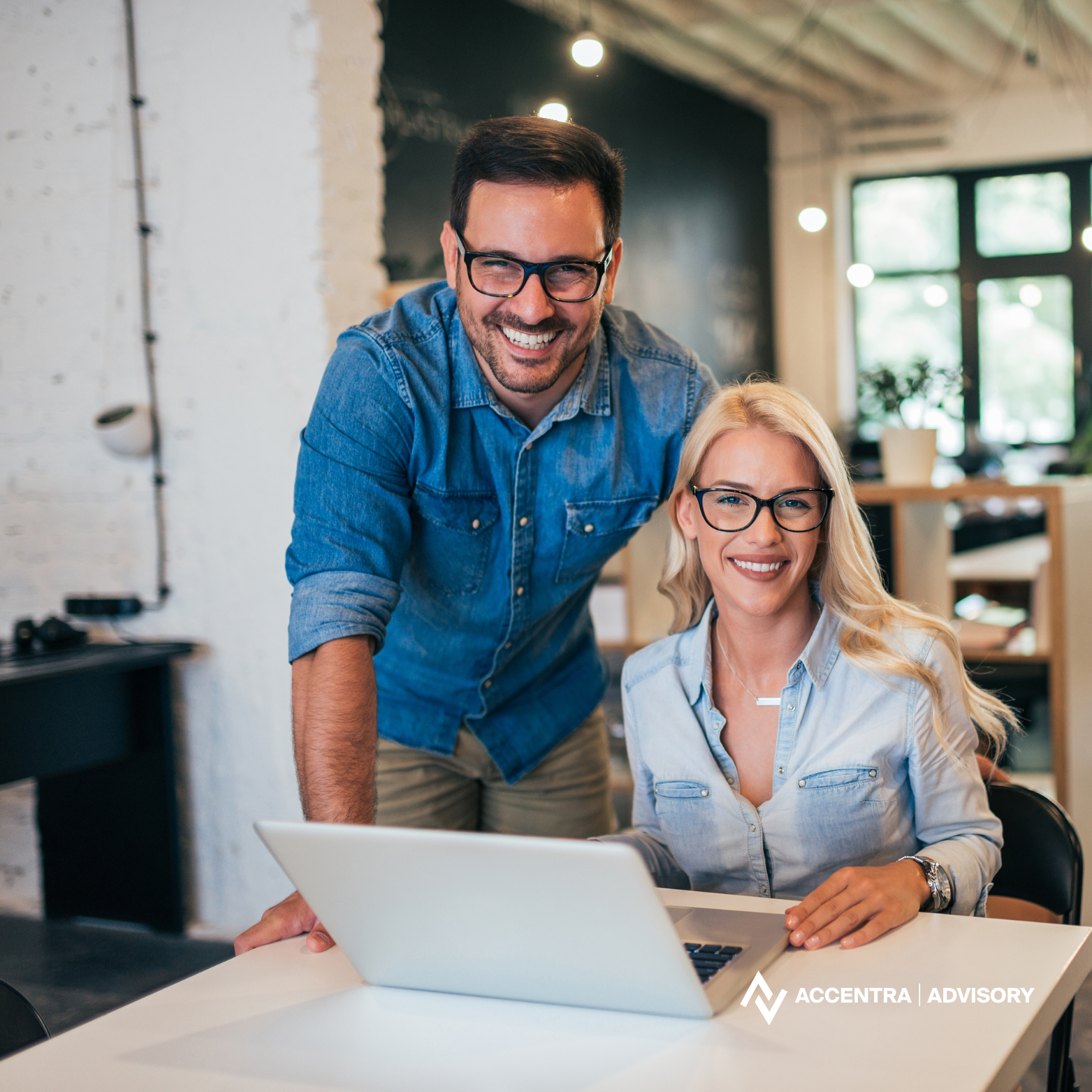 A man and woman smiling at a laptop in a modern office space.