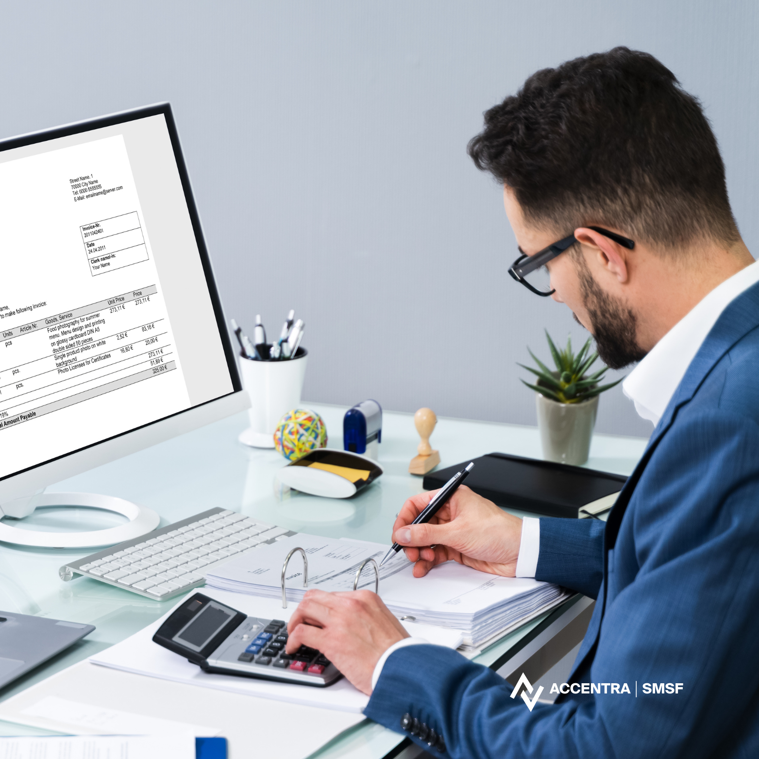 Man in blue suit working at a white desk with a calculator, papers, and a computer monitor displaying an invoice; desk also has office supplies, a small plant, and a computer keyboard.