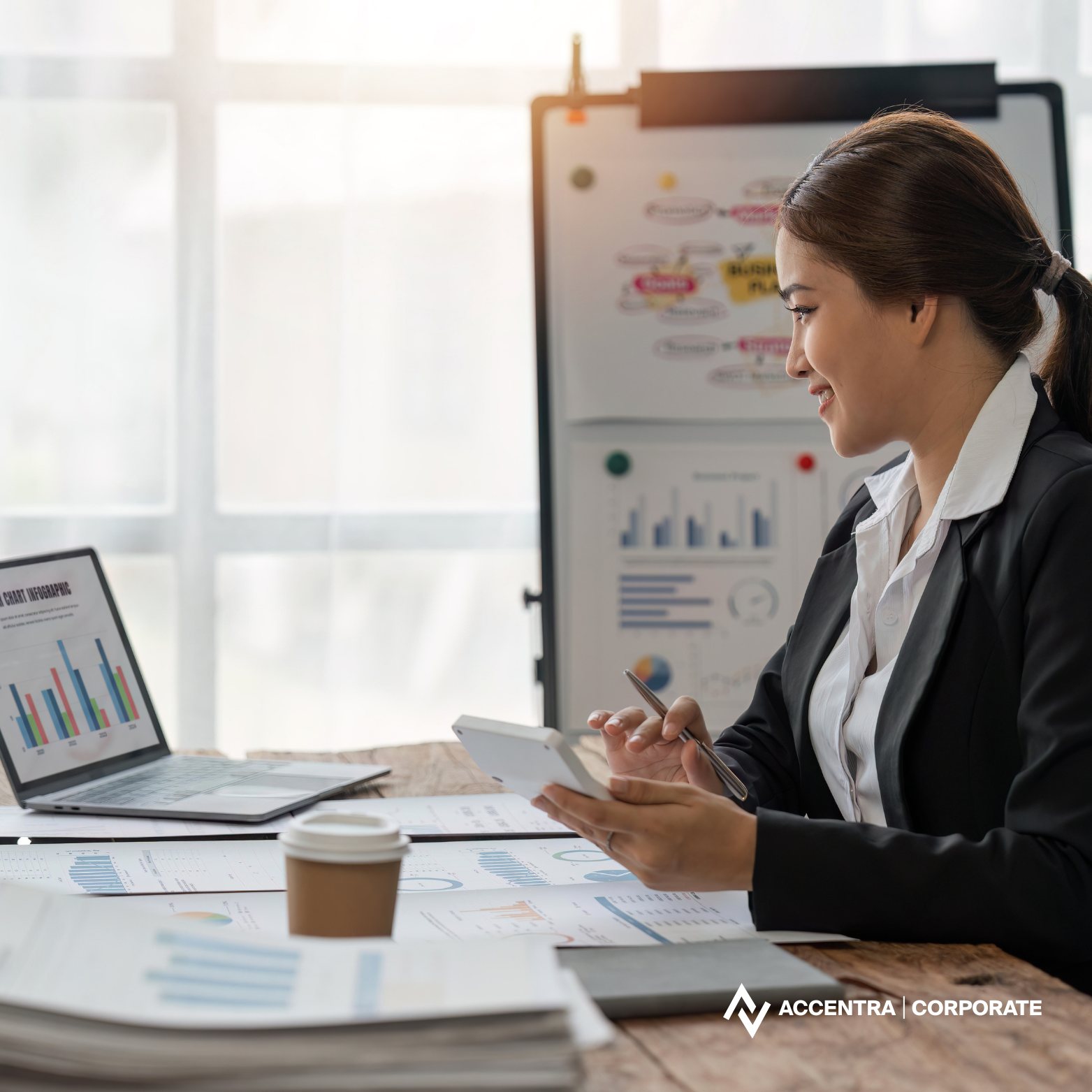 Businesswoman working at desk with charts and graphs, using smartphone, in office.