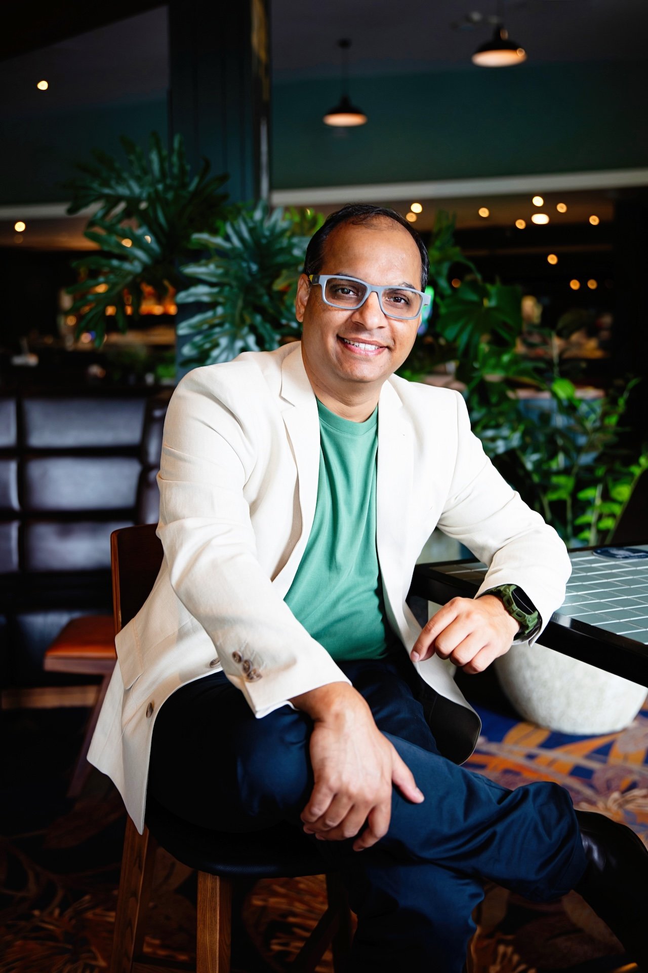 A man wearing glasses, a cream blazer, and a green t-shirt, sitting in a restaurant or cafe with green plants and warm lighting, smiling at the camera.