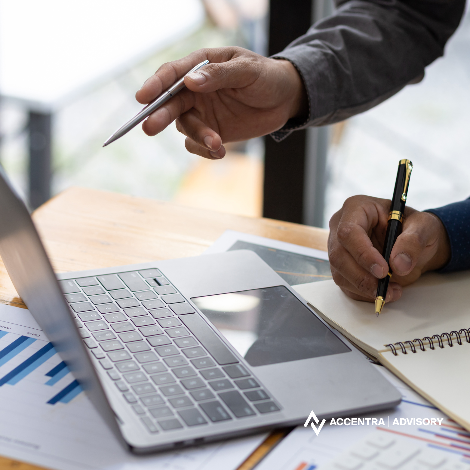 Two people are working at a desk with a laptop, papers, and notebooks, one is holding a pen and the other is holding a silver pen.