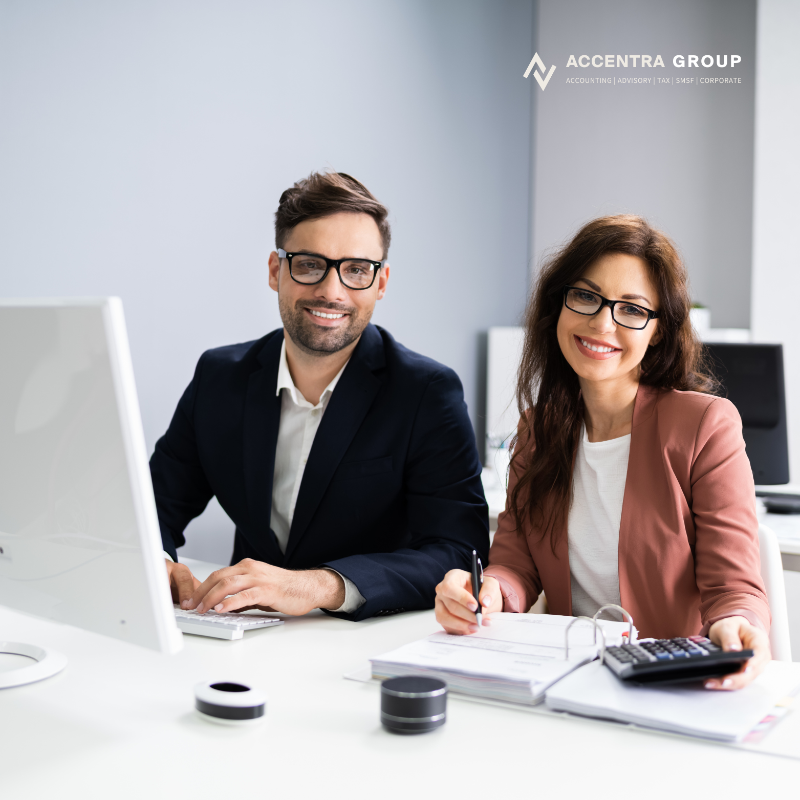 Two professionals, a man and a woman, sitting at a desk in a modern office, smiling and looking at the camera. The woman is holding a calculator and a pen, with notebooks in front of her. The man is typing on a computer keyboard. The background features a desk with a monitor and a wall with the Acctera Group logo.