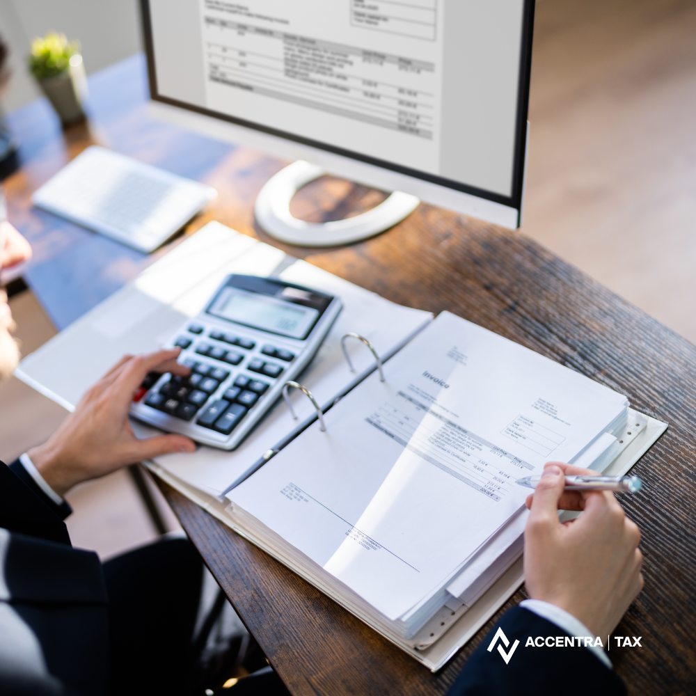 A person working at a desk with an open binder, calculator, and pen, while looking at a computer monitor displaying a document. There are office supplies and a small plant on the desk.