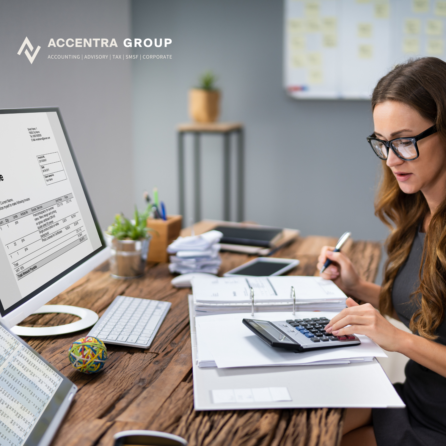 Woman working at a cluttered wooden desk with a computer, calculator, and documents, in an office with a whiteboard and potted plant in the background.