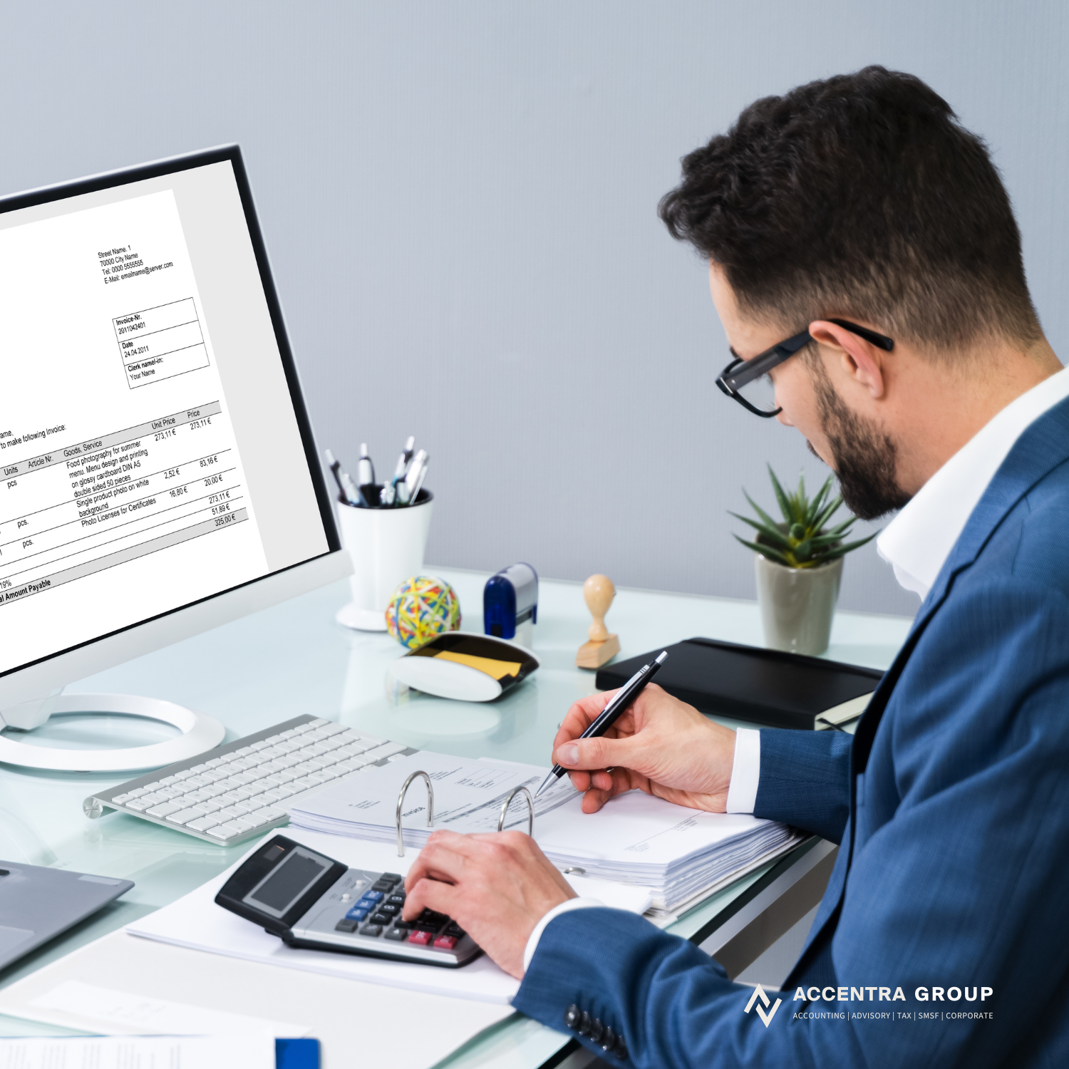 A man in a blue suit and glasses sitting at a desk, working with a calculator, pen, and documents, with a computer monitor displaying an invoice. The desk has a white cup with pens, a small plant, and office supplies.