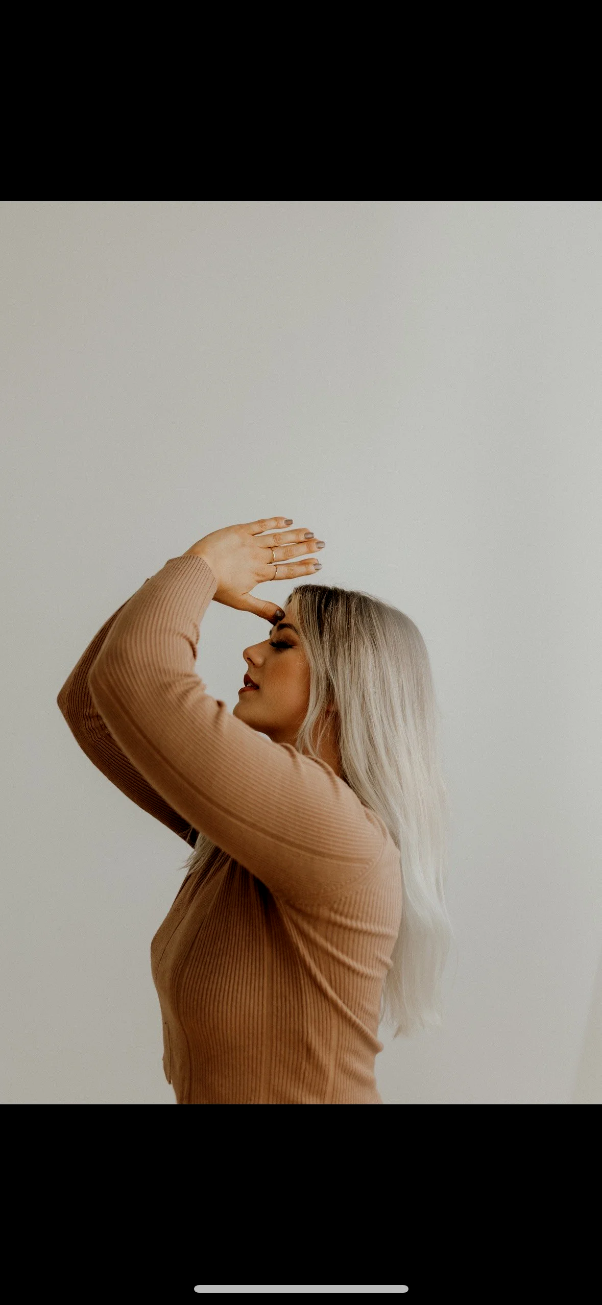 A woman with long blonde hair, wearing a light brown ribbed shirt, stands with her eyes closed and one hand on her forehead, posing against a plain white background.