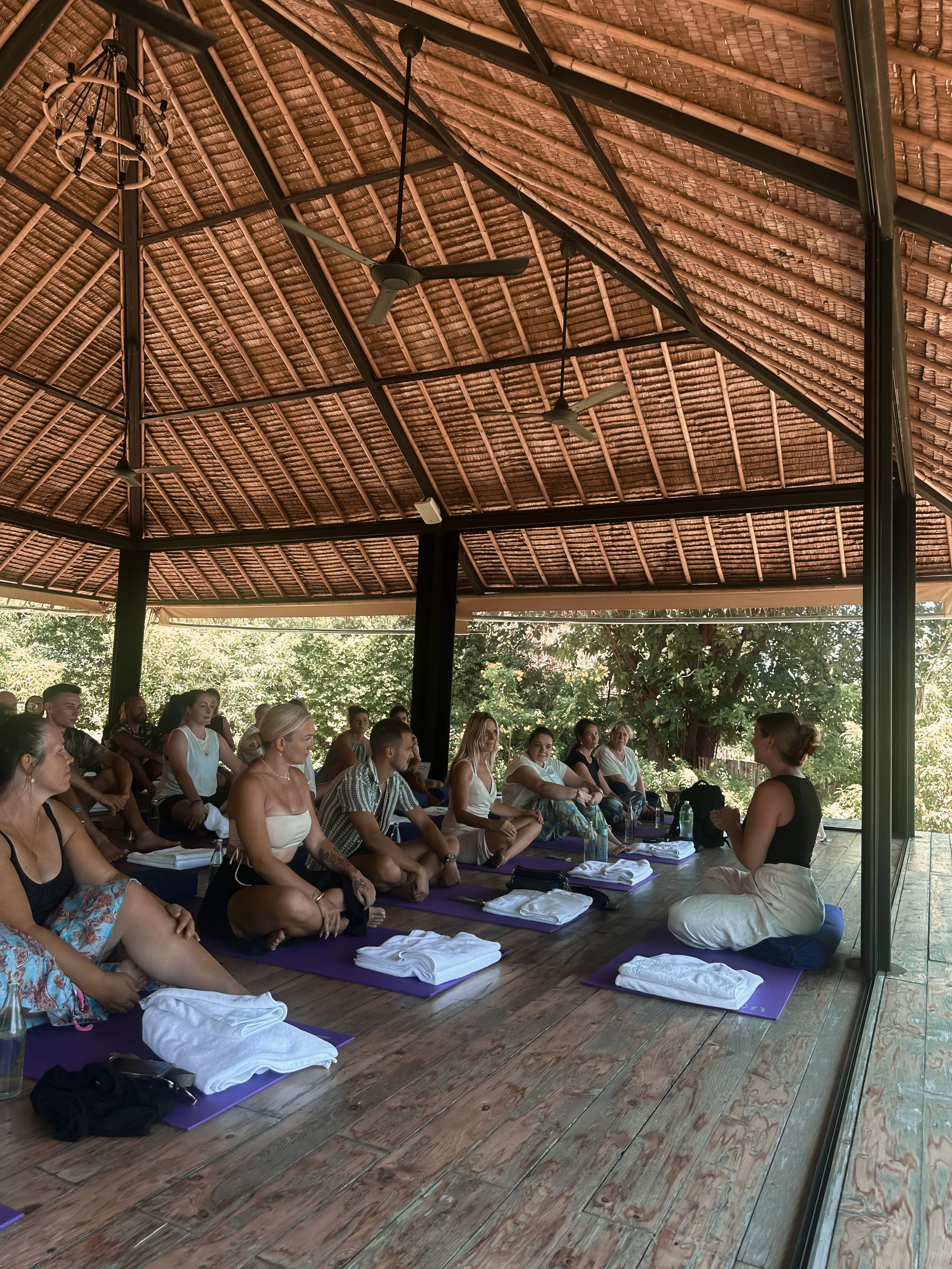 A group of people participating in a yoga class or workshop on purple mats under a thatched roof with ceiling fans, with a female instructor at the front.