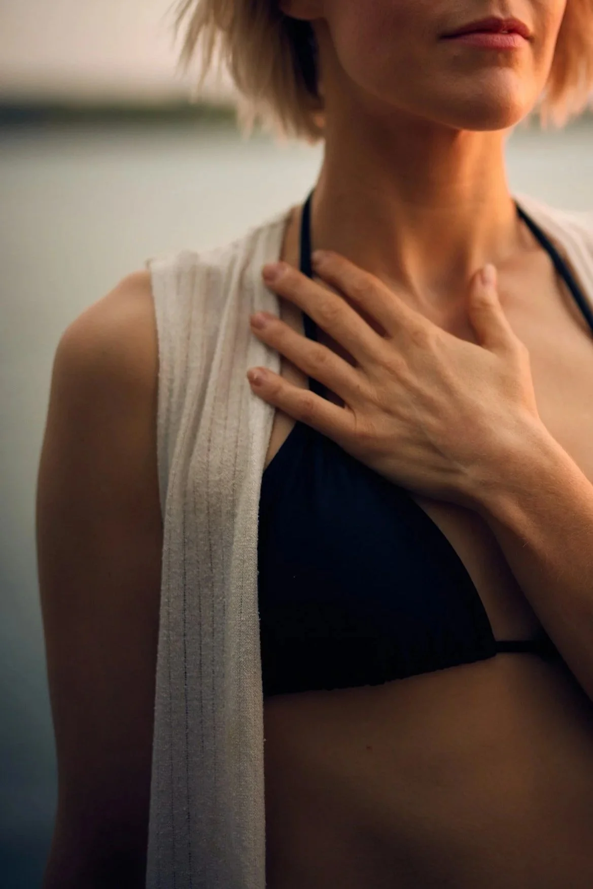 A woman with short blonde hair, wearing a black bra and a white towel over her shoulder, is touching her throat with her right hand, standing near a body of water.