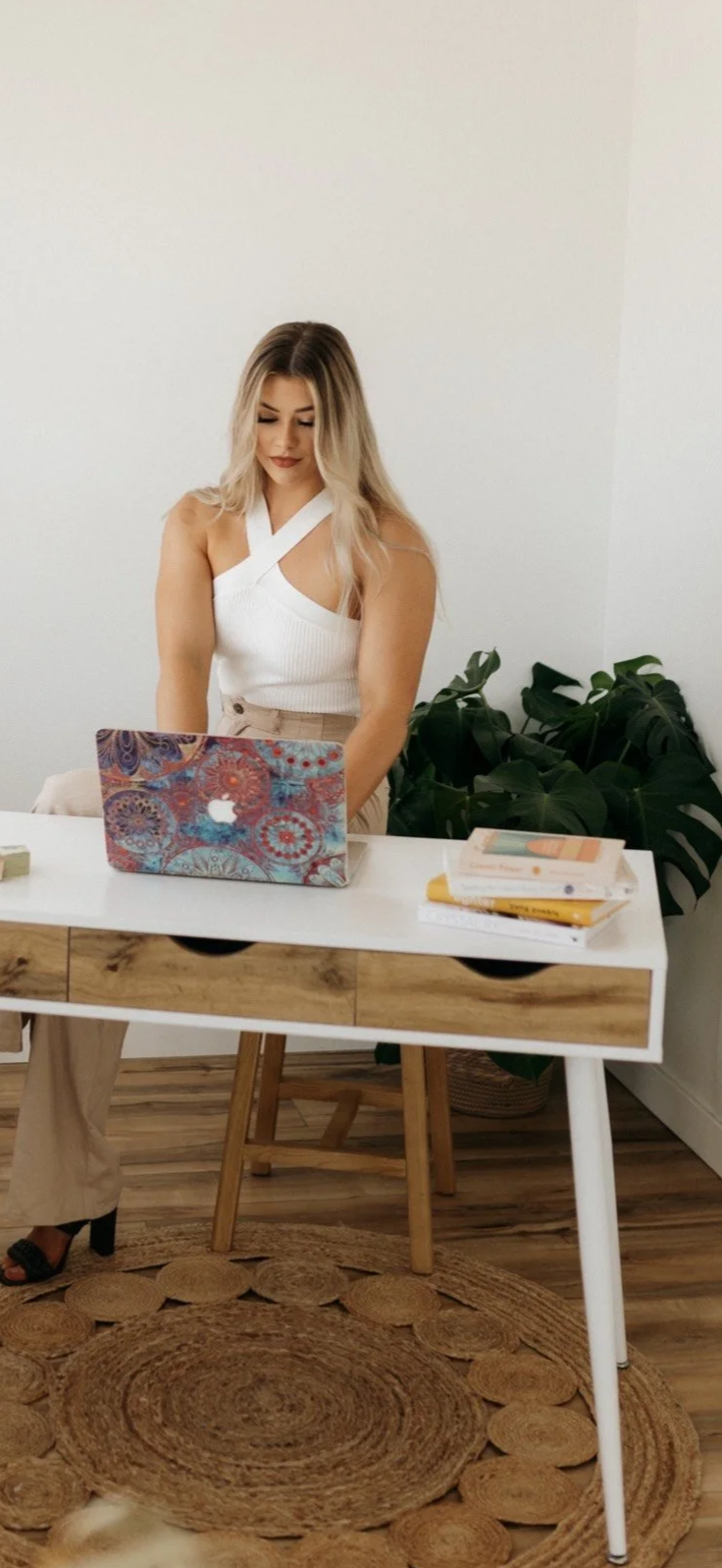 A woman with long blonde hair working on a colorful laptop at a white desk, in a room with a white wall, wooden floor, and a large green plant in the background.