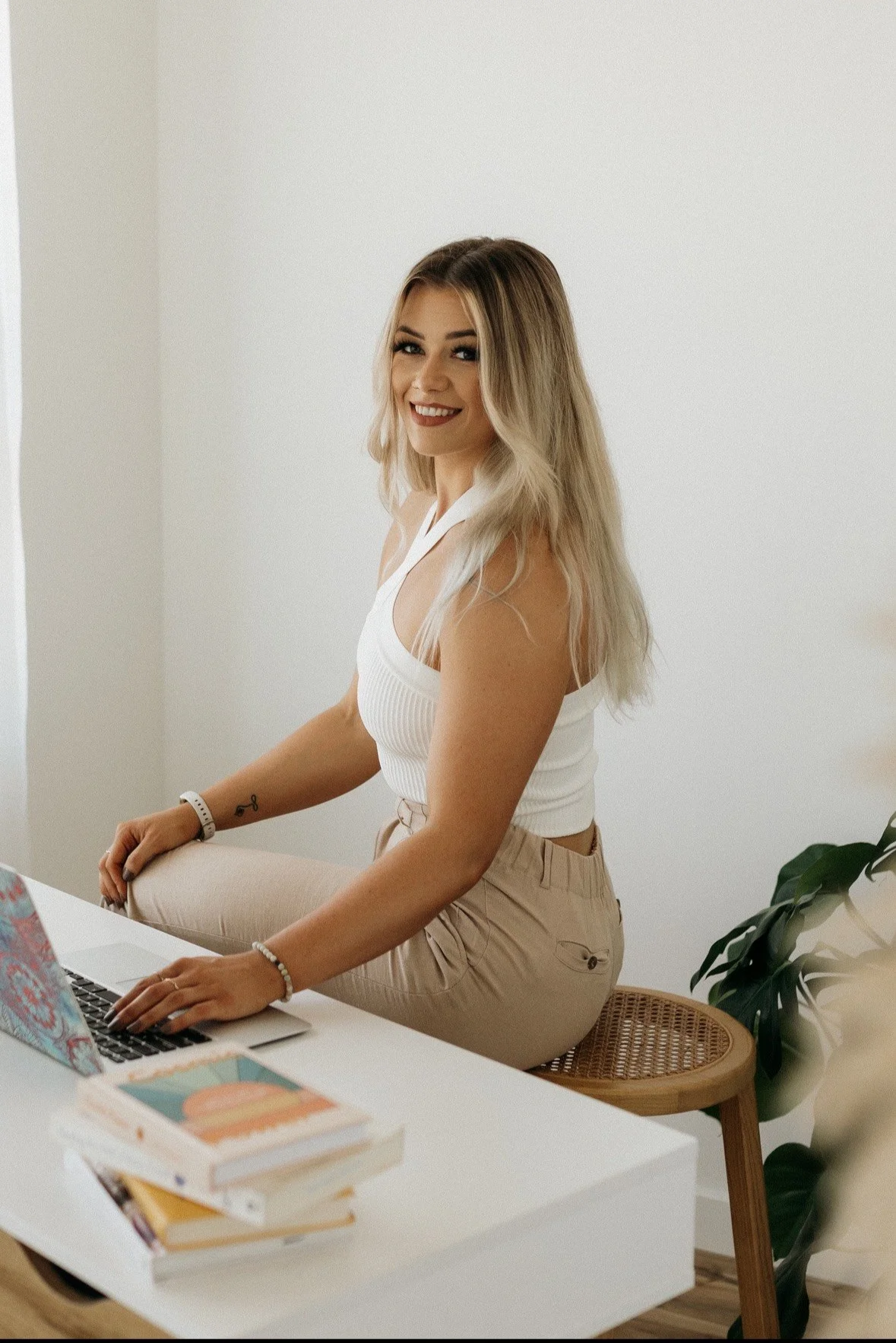 A woman with long blonde hair sitting at a desk, smiling at the camera, wearing a white sleeveless top and beige pants, with a laptop, books, and a plant nearby.