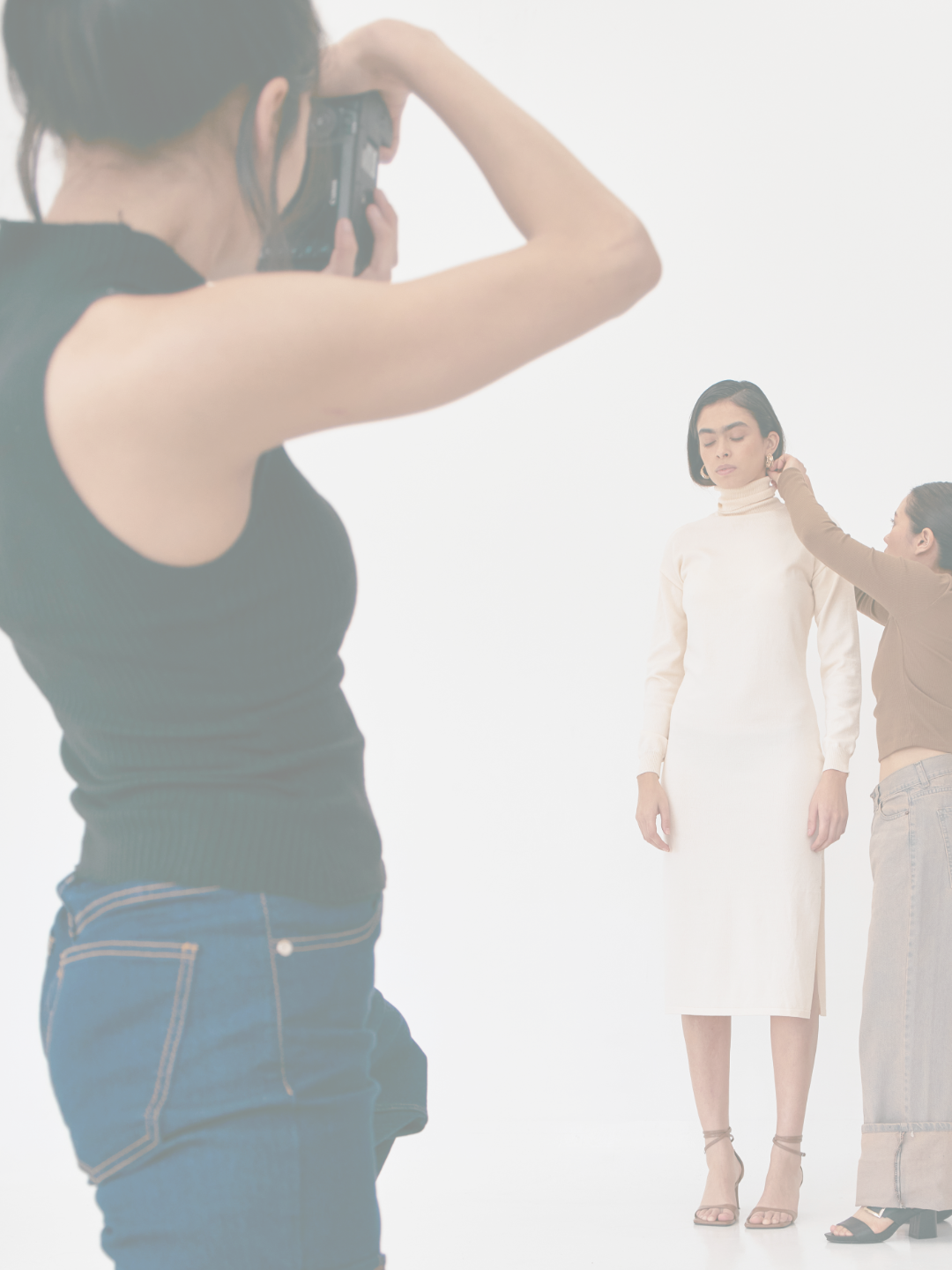 Photo of a woman model being styled during a photoshoot, with a photographer taking picture and a stylist adjusting the model's earring.