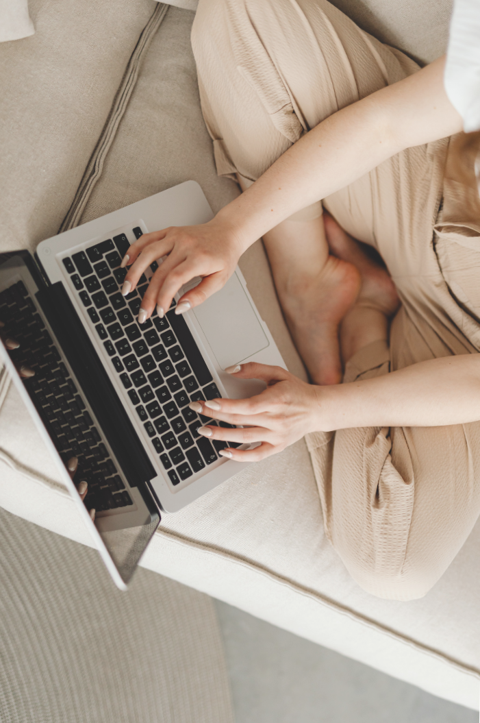 Person sitting on a beige couch using a silver laptop, with legs crossed and hands on the keyboard.