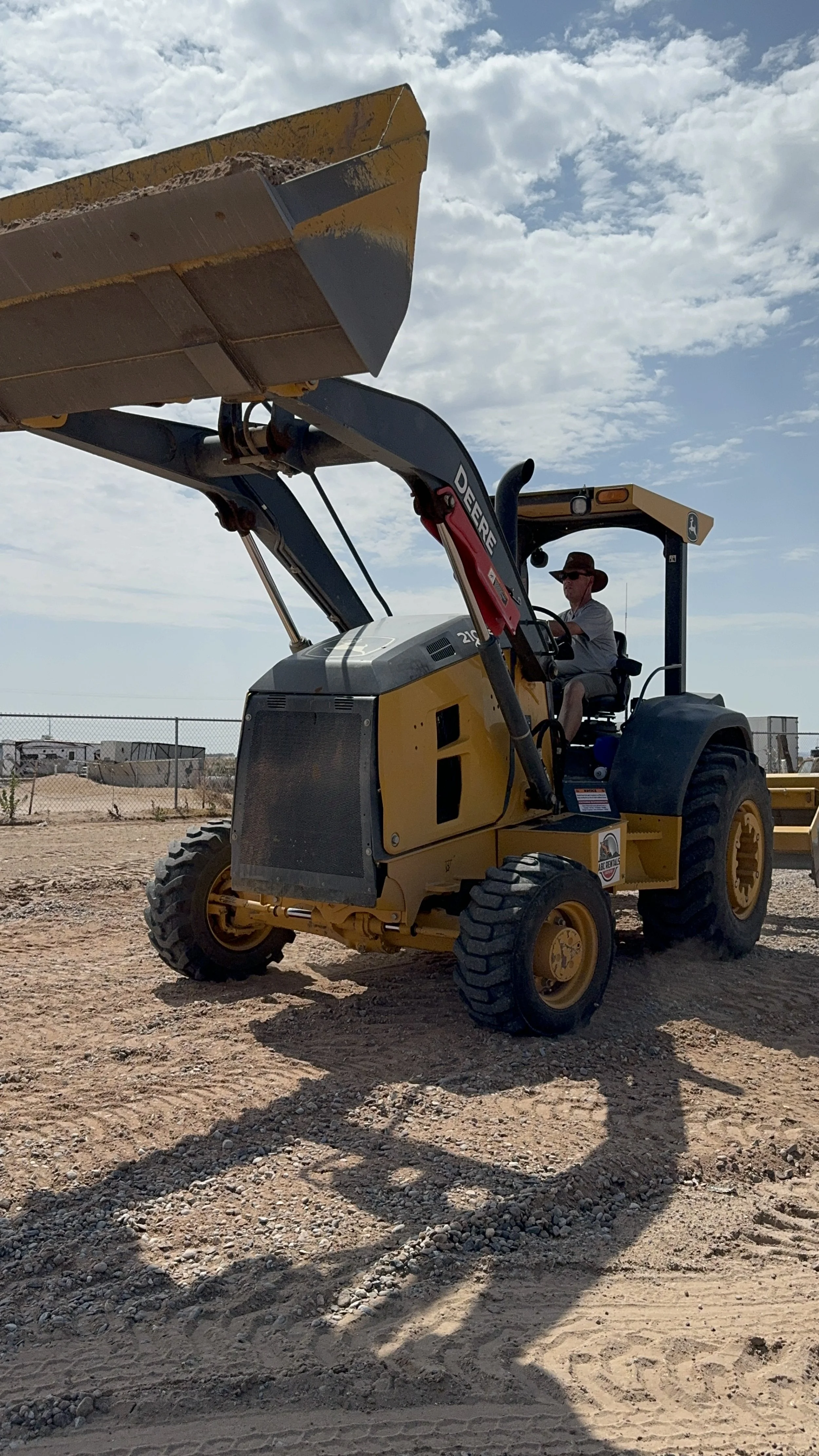 A man wearing a hat and sunglasses operating a yellow John Deere backhoe loader on a construction site with a chain-link fence and trailers in the background, under a partly cloudy sky. Building a road for Stanfield Clinic, Stanfield, Arizona.