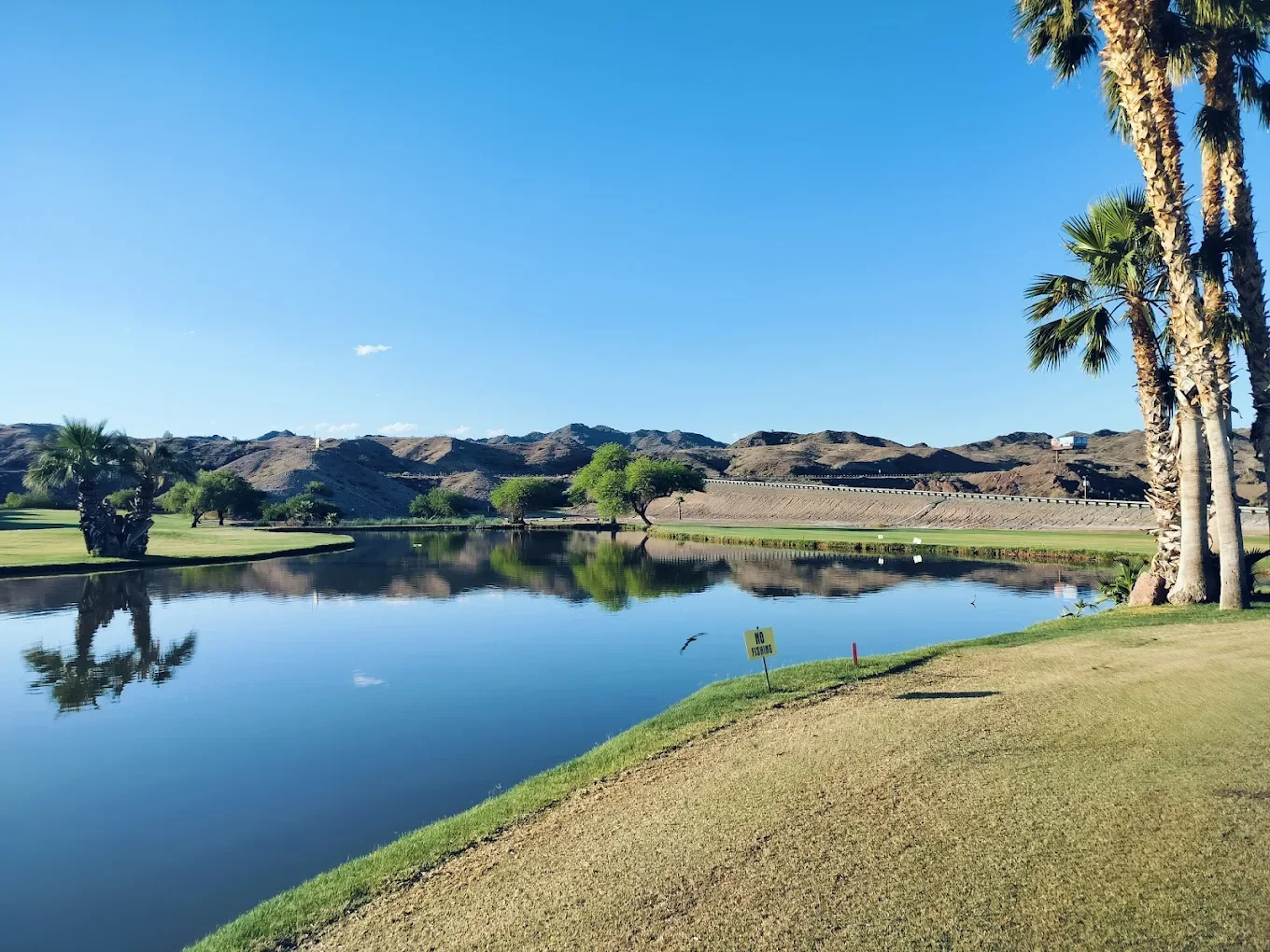 A scenic landscape with a calm river reflecting the clear blue sky, palm trees, and distant hills, with a grassy area in the foreground and a yellow sign that reads 'No Fishing'. Endangered Species ponds. Emerald Canyon Golf Course. La Paz County, AZ