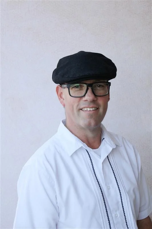 Dr. Daniel Barbara, Partner and Principal Consultant of Pacific Rim EcoSystemic Strategy Advisors wearing a black hat, black glasses, white shirt with decorative black stitching, standing against a plain light-colored background.