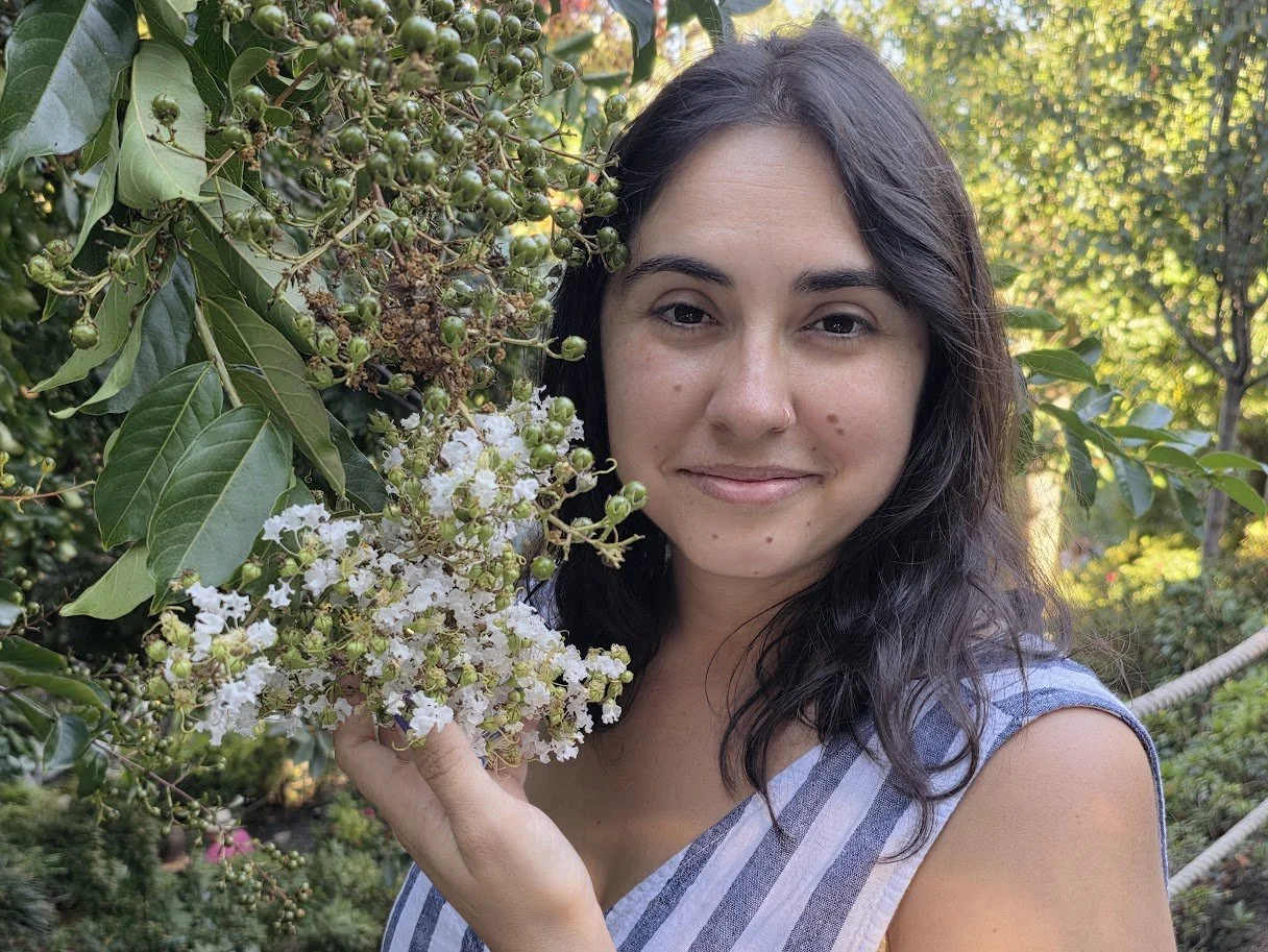 Erika Vega, Partner and Senior Consultant of Pacific Rim EcoSystemic Strategy Advisors with dark hair and a light complexion holding a branch with white flowers and green berries, smiling outdoors in a garden.