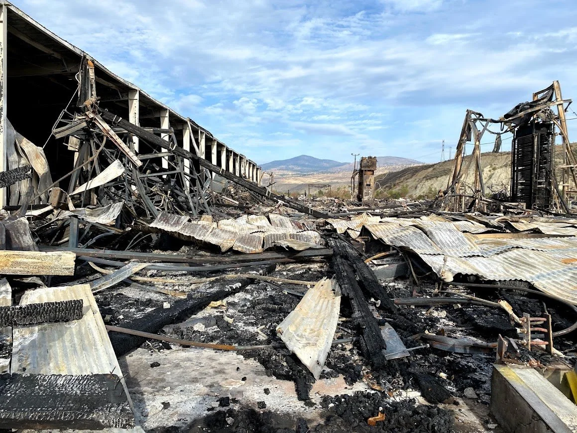 Burned and destroyed industrial building with metal debris and burnt roofing, landscape with hills and cloudy sky in the background. Omak, Washington, Brownfields site.