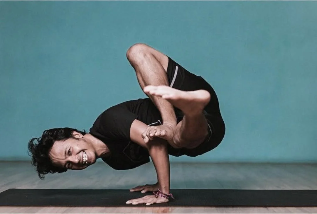 A person performing a complex yoga pose on a black mat in front of a plain turquoise background.