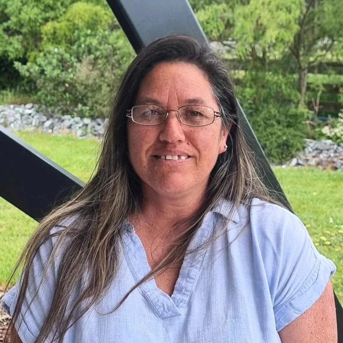 A woman with long hair, glasses, and a light blue shirt, smiling outdoors in front of green trees and grass.