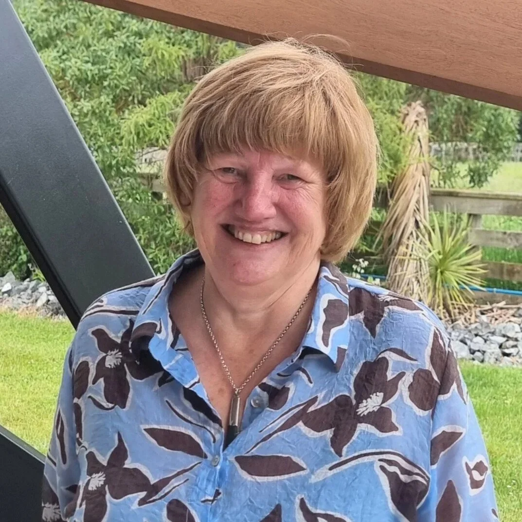 A smiling middle-aged woman with short, light brown hair, wearing a blue floral shirt and a silver necklace, outdoors with green bushes, grass, a wooden fence, and a patio umbrella in the background.