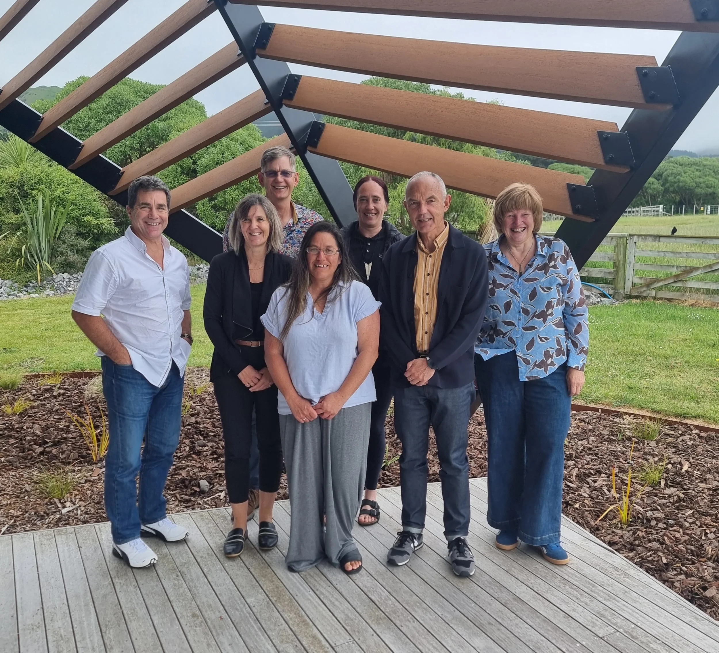 Group of eight people standing outdoors on a wooden deck, smiling, with a modern wooden and black metal structure behind them, green trees, and a fenced field in the background.