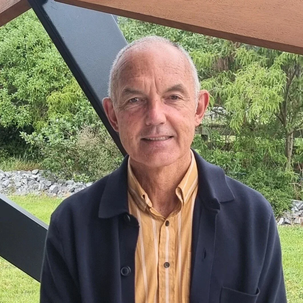 A smiling elderly man with short gray hair, wearing a yellow striped button-up shirt and a dark blazer, standing outdoors with green trees and grass in the background.