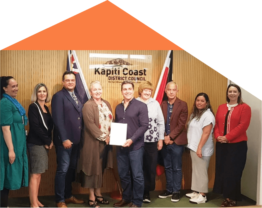 Group of nine people standing in front of a wooden wall with a 'Kapiti Coast District Council' sign, two flags, and a man holding a document.