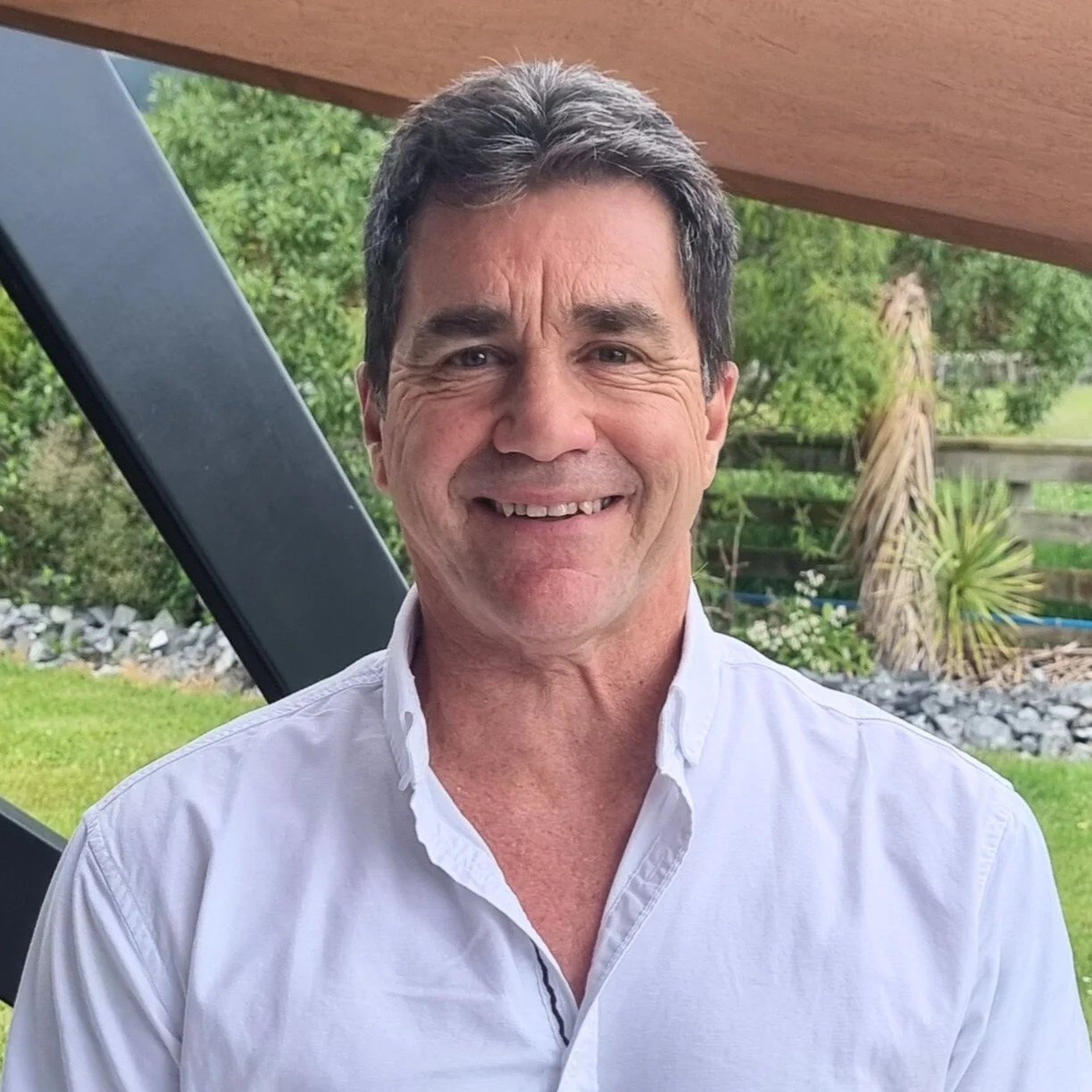 A middle-aged man with dark hair and a white shirt smiling outdoors with trees and plants in the background.