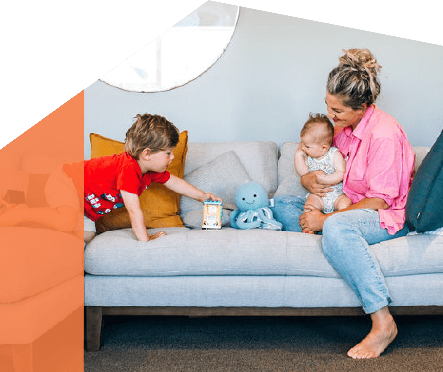 A woman in a pink shirt sitting on a gray couch holding a baby while a young boy in a red shirt reaching for an octopus toy on the couch.