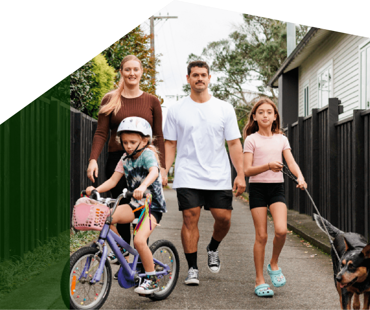 A family of five walking down a sidewalk with a young girl riding a bike, a woman, a man, a girl holding a dog leash, and a dog walking beside them.