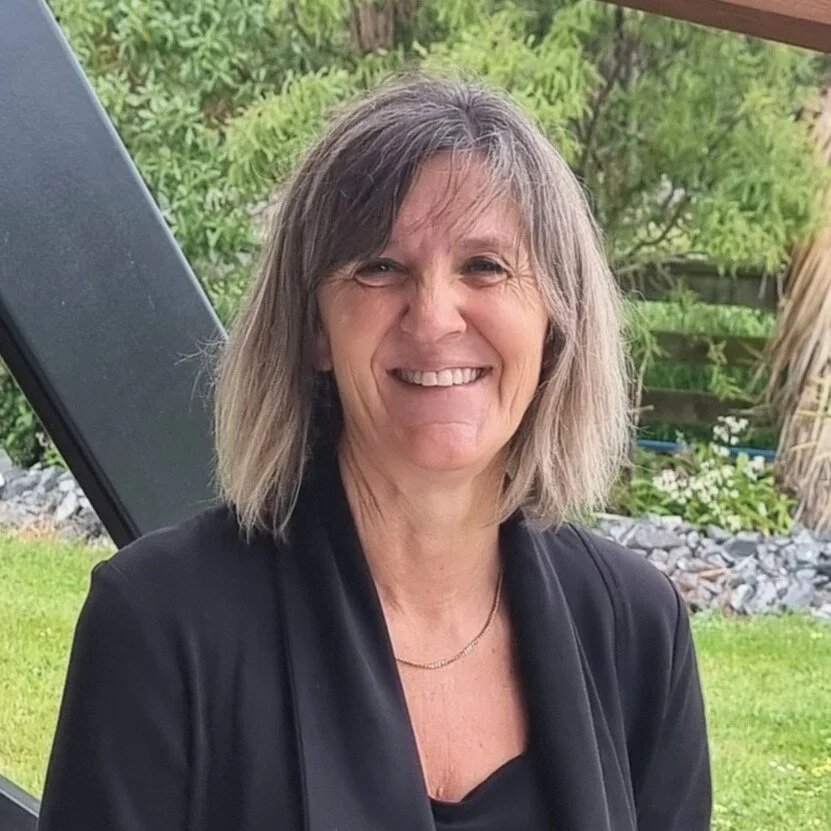 A woman with shoulder-length gray hair, smiling and wearing a black jacket, standing outdoors with greenery in the background.
