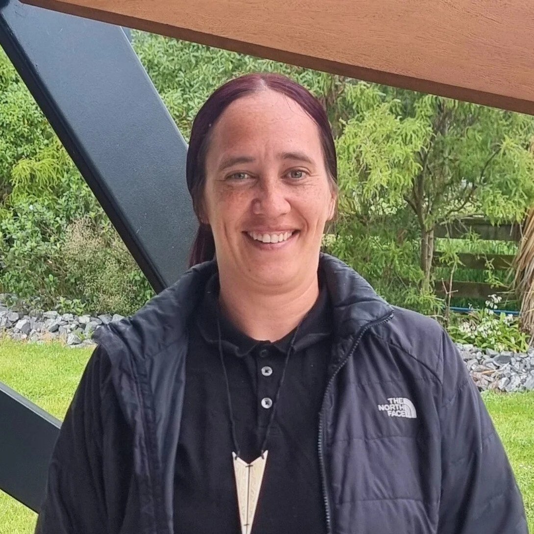 Smiling woman with dark brown hair wearing a black The North Face jacket and a black shirt, standing outdoors with greenery in the background.