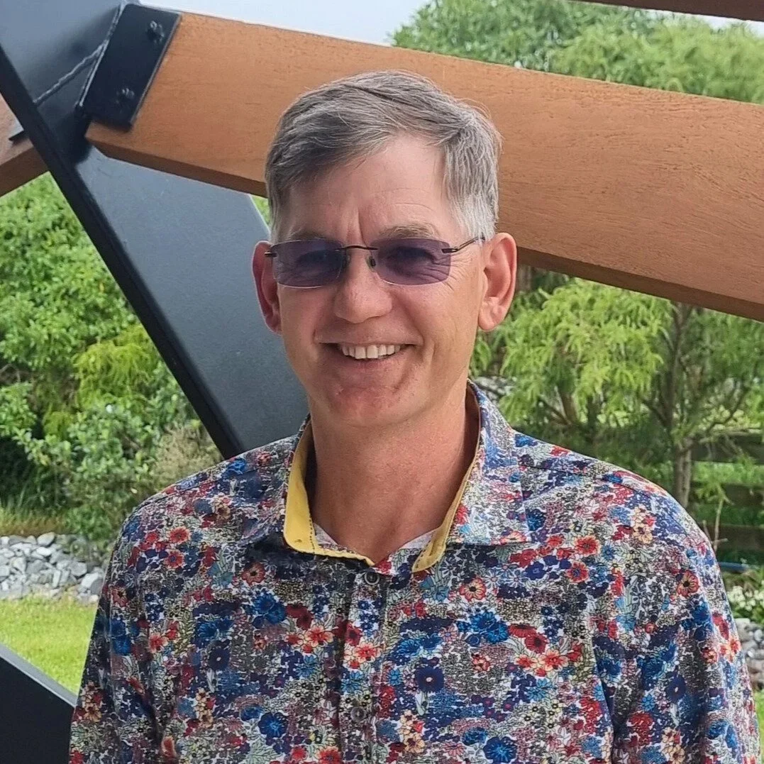 A smiling man with gray hair and glasses, wearing a colorful floral shirt, standing outdoors with green trees and a garden in the background.