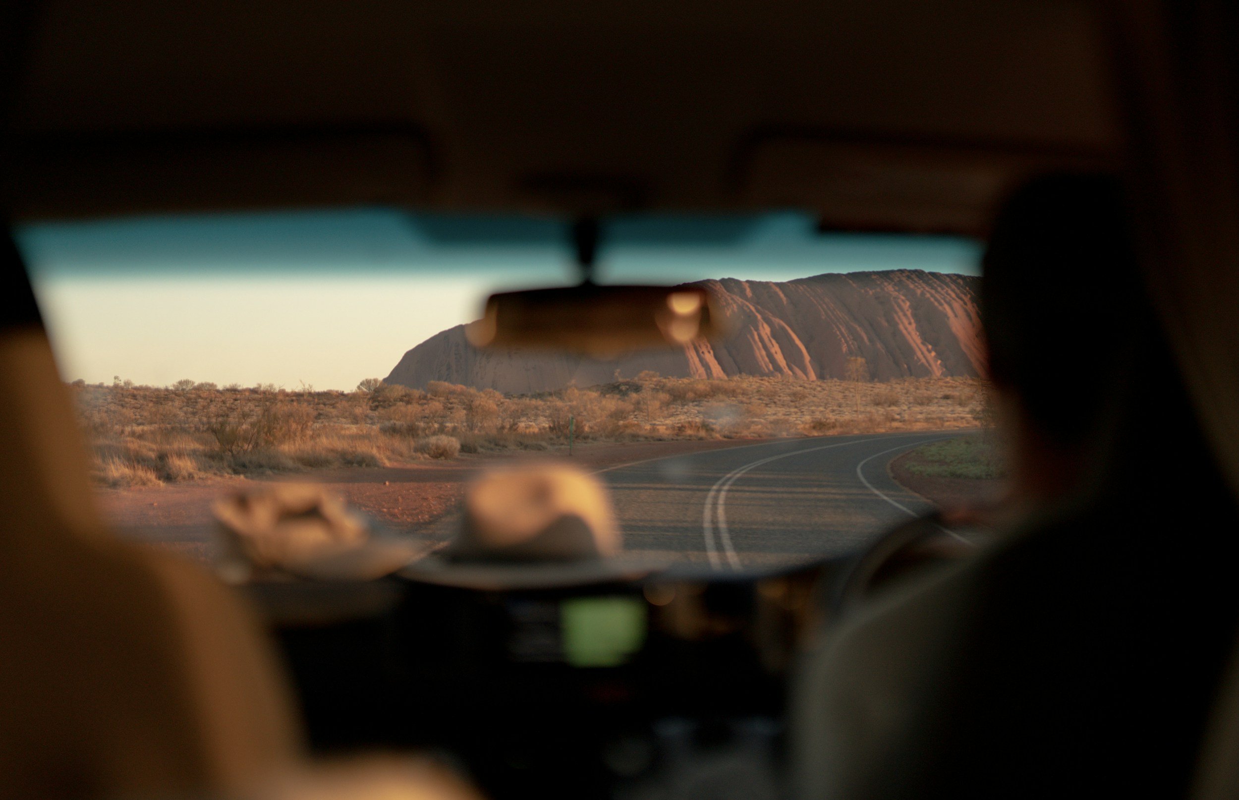 View of Uluru rock formation seen through the rearview mirror of a vehicle during sunset or sunrise in the desert.