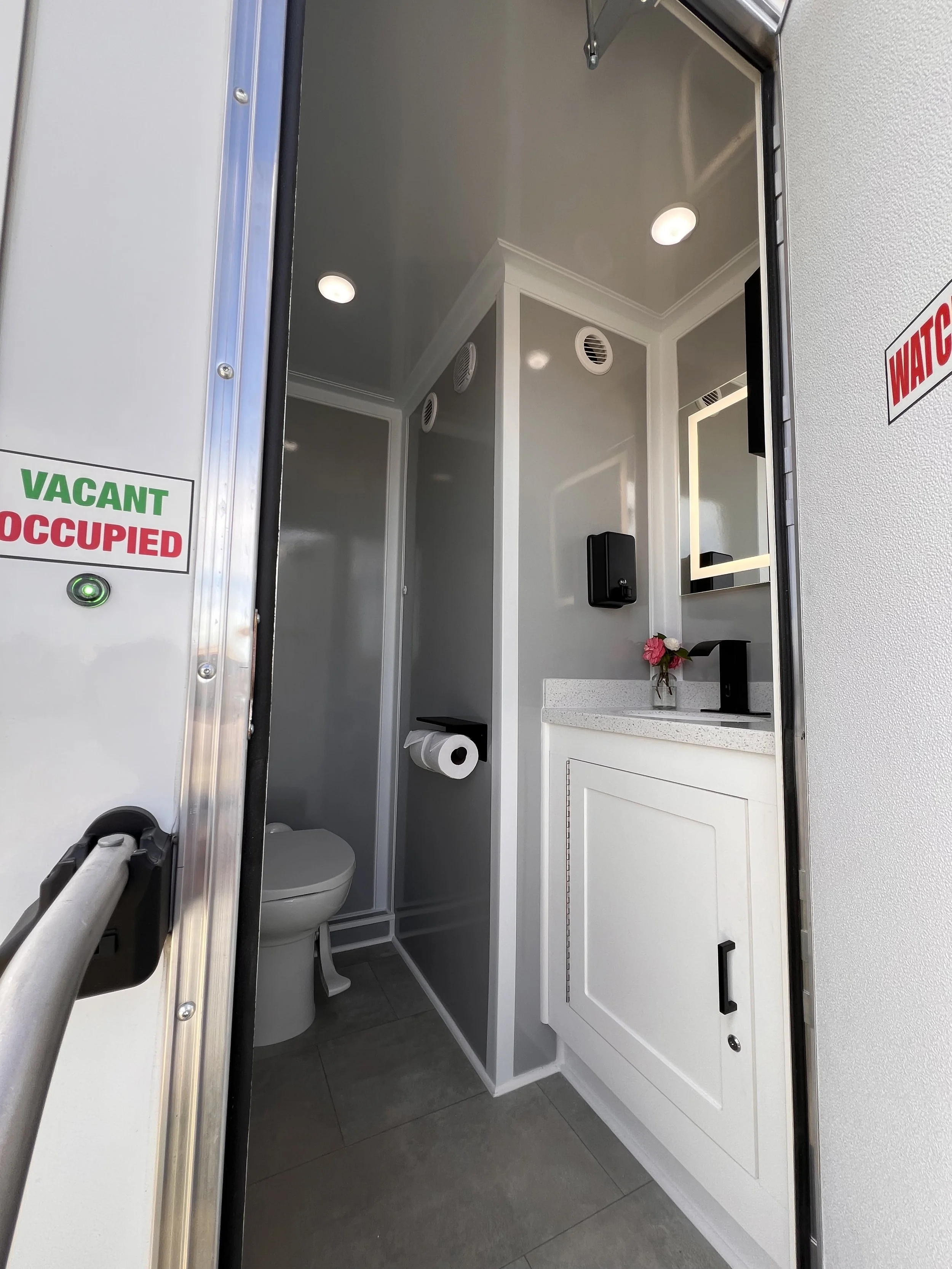 A modern bathroom with a white vanity, a mirror with LED lighting, a black faucet, a wall-mounted black tissue dispenser, and a toilet in the foreground. The bathroom features gray walls, a dark gray door, and gray tile flooring.