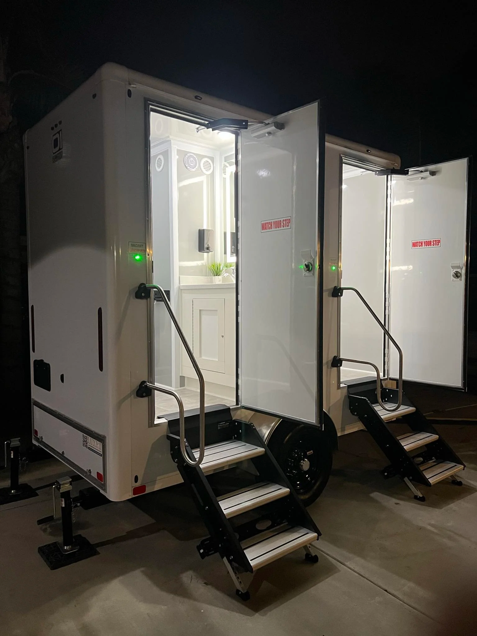White trailer with two stairs leading to separate restroom doors, one for women and one for men, located outdoors at sunset with palm trees in the background.