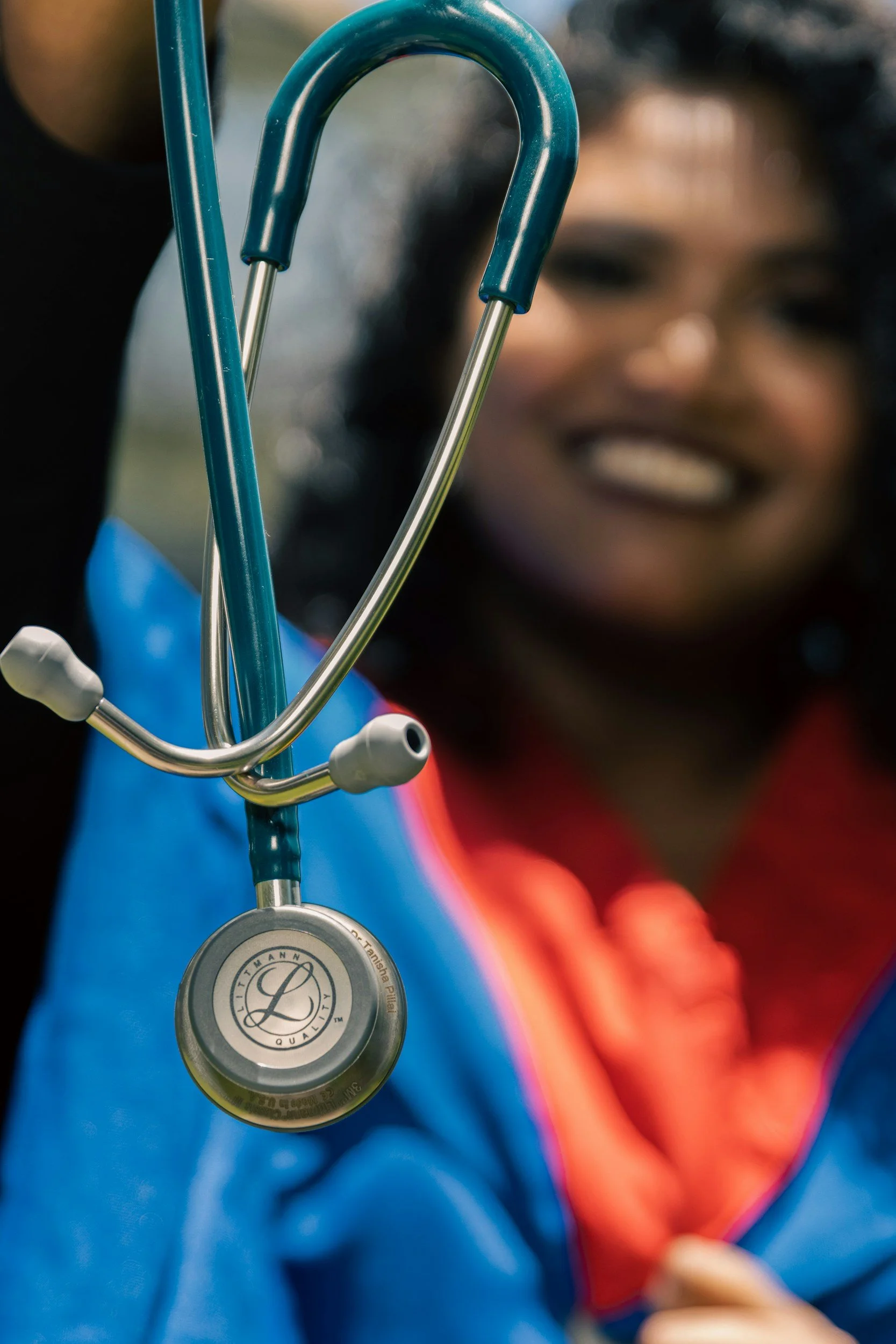 A healthcare worker in a blue uniform holding a stethoscope with a smiling face in the background.