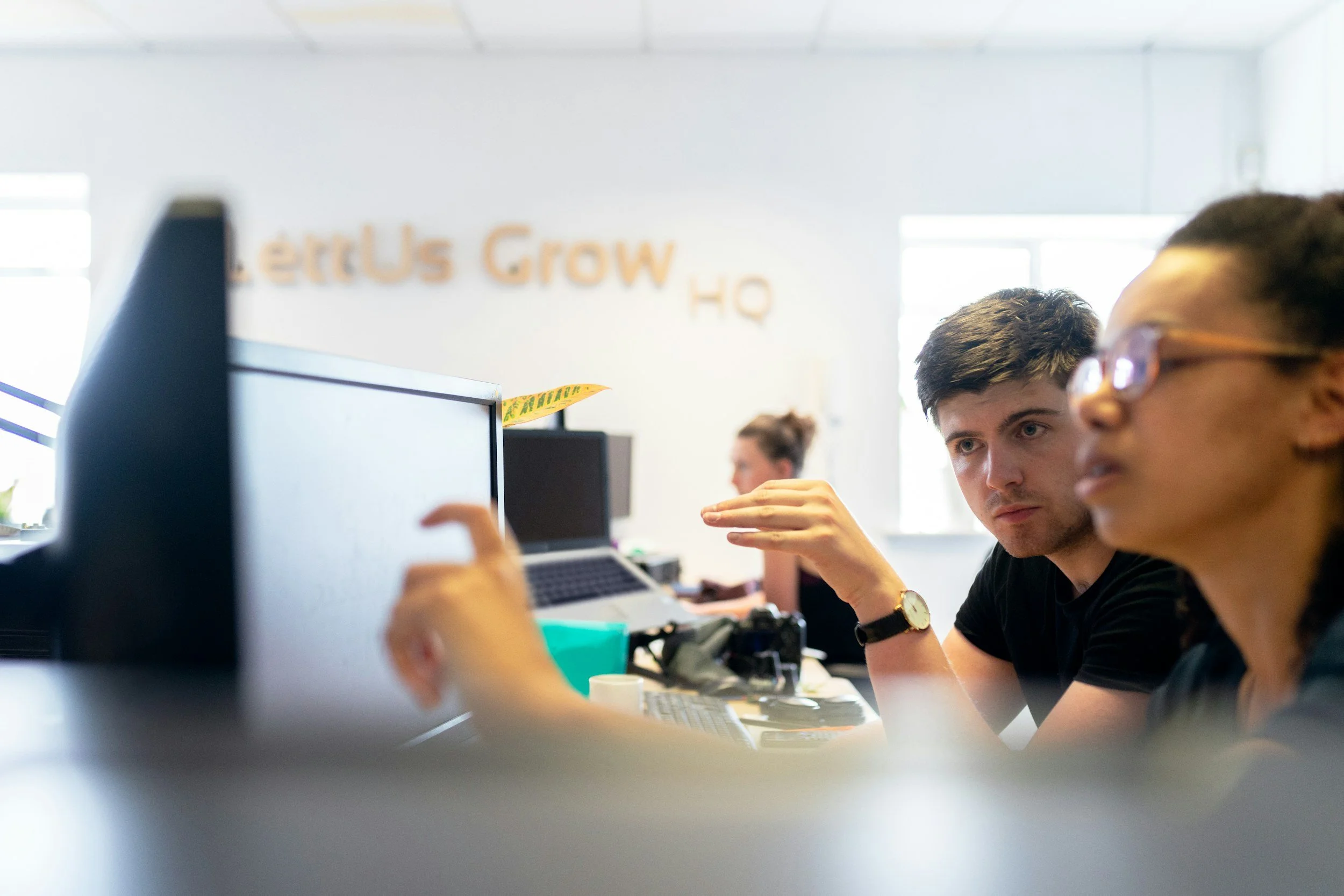 Two people working at a desk in an office, with a woman on the right and a man on the left, focusing on a computer screen, with office chairs, laptops, and a blurred woman in the background. The wall in the background has the text 'LetUs Grow HQ'.