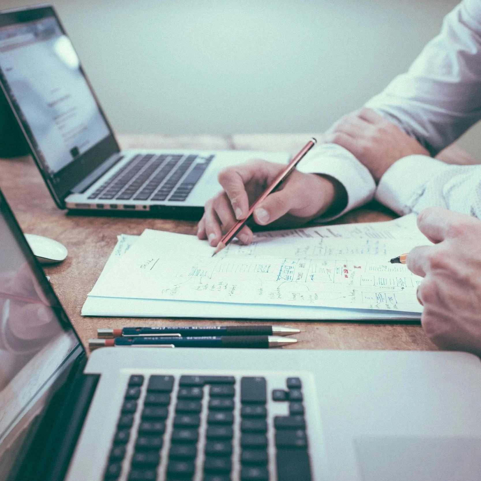 Two people working at a desk with laptops, notebooks, and pens, engaged in a collaborative work session.