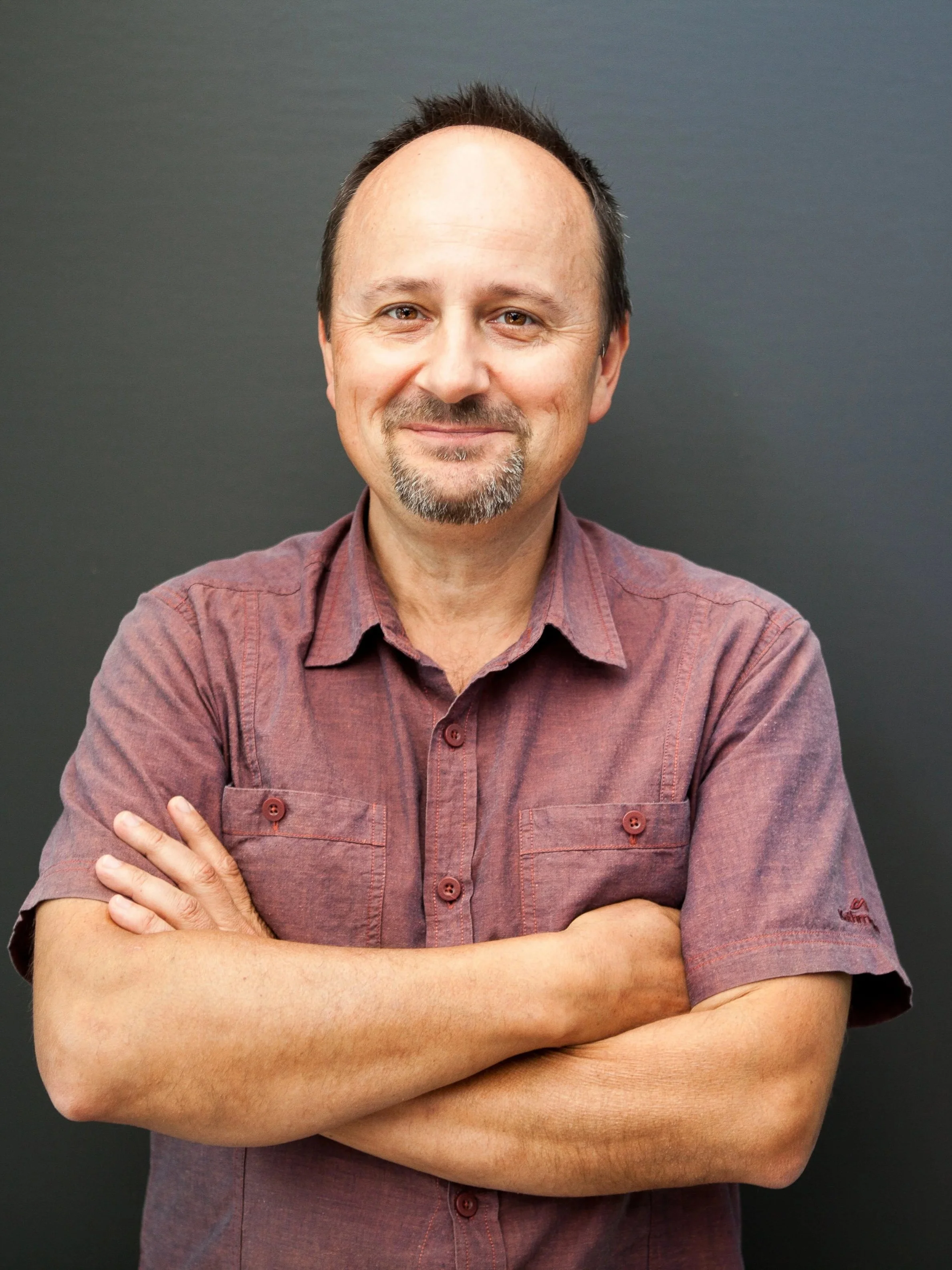 A middle-aged man with short dark hair and a goatee, smiling with arms crossed, wearing a short-sleeved maroon shirt, standing against a dark background.