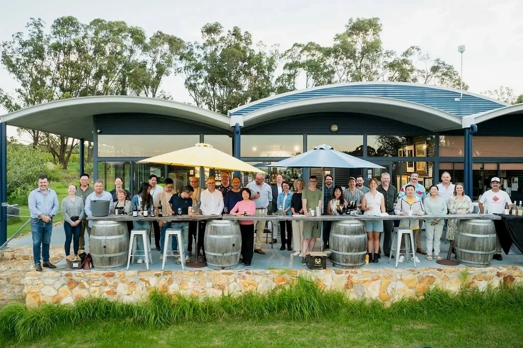 Group of people gathered outdoors at a building with a modern curved roof, standing behind a long bar made of wine barrels, with umbrellas providing shade, during daytime.