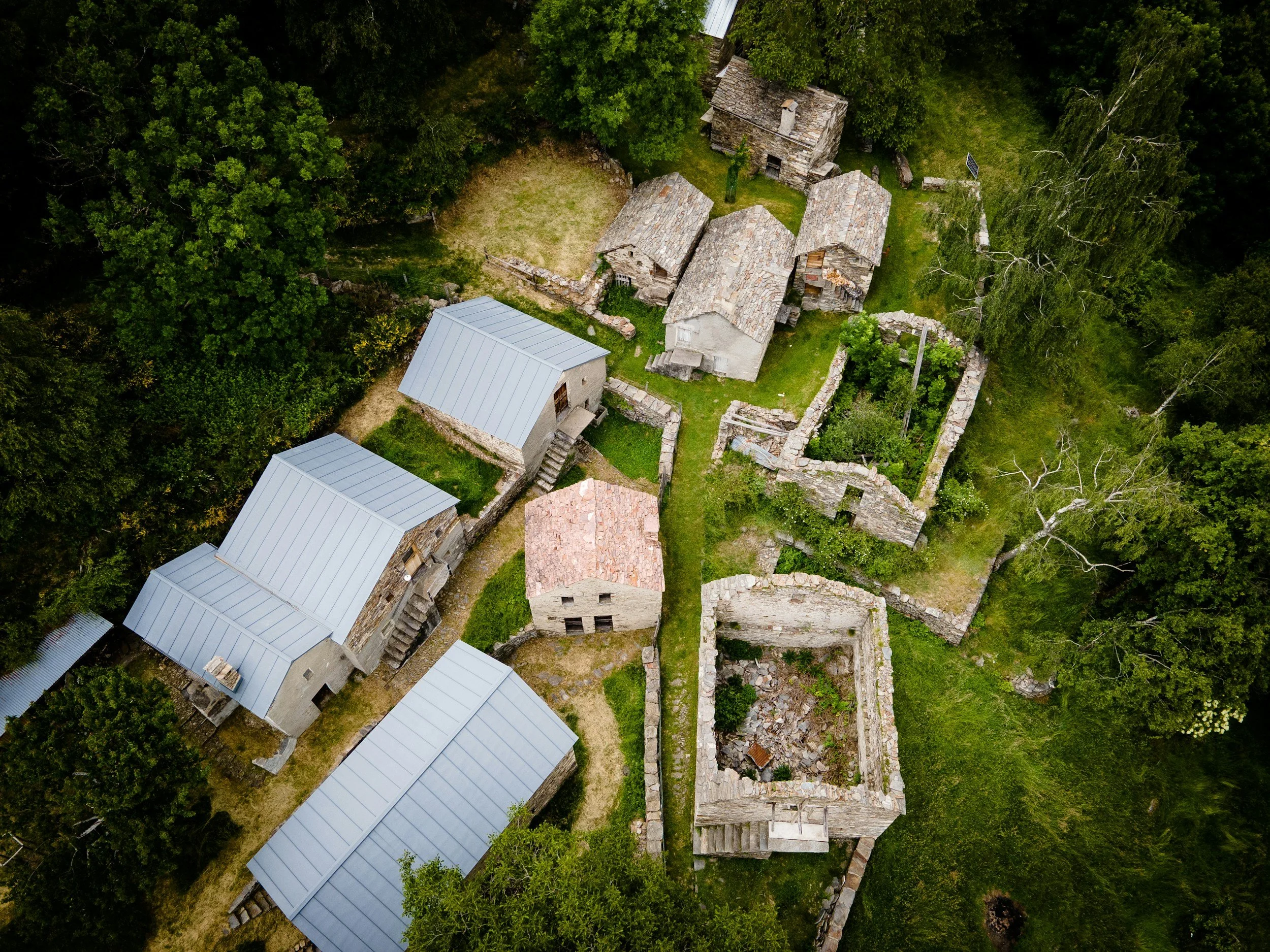 Aerial view of an abandoned stone village with several stone buildings, some with metal roofs, surrounded by dense green trees and grass.