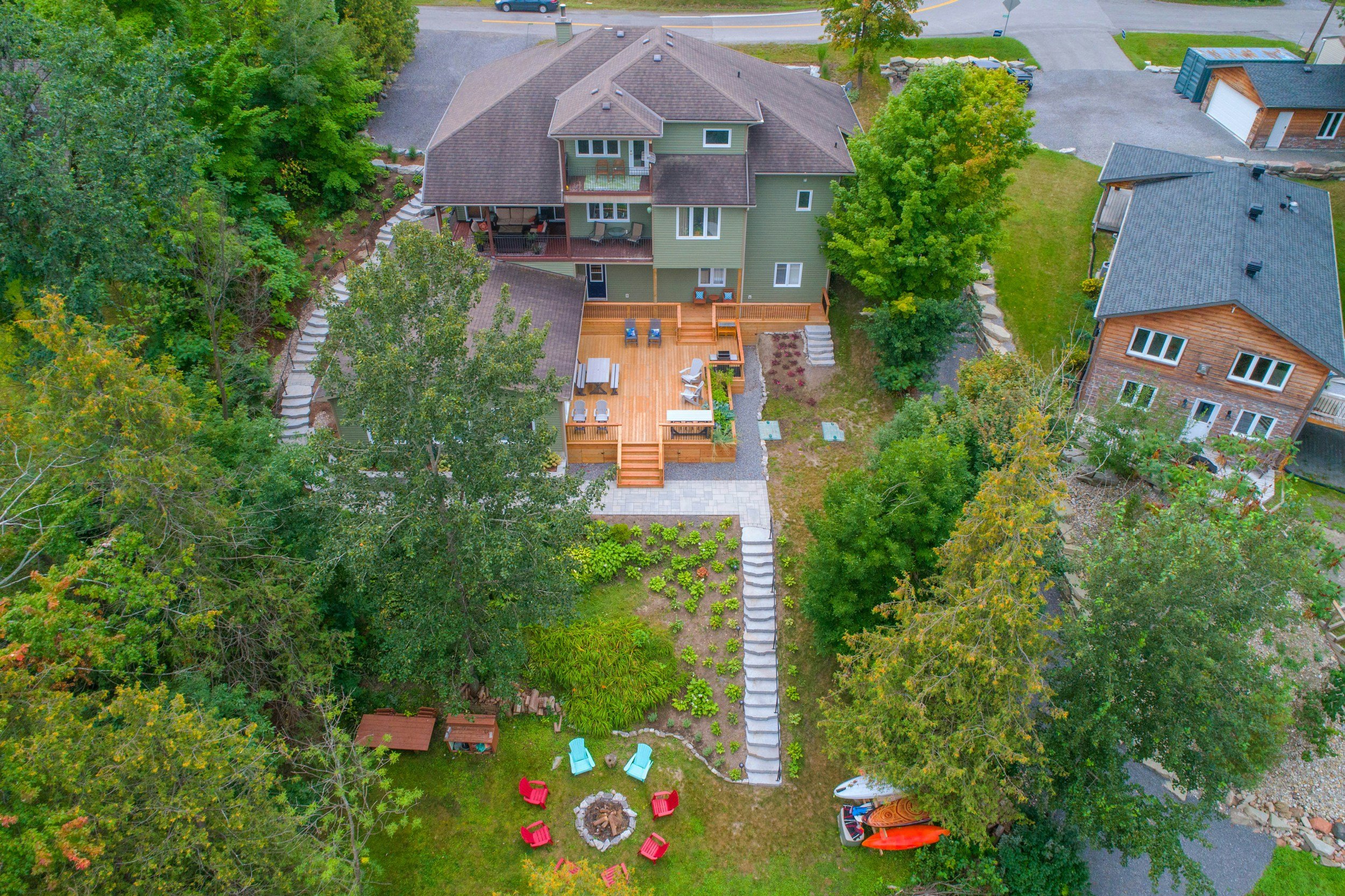 An aerial view of a backyard with a large wooden deck, outdoor furniture, a fire pit surrounded by colorful chairs, and lush green trees around the yard.