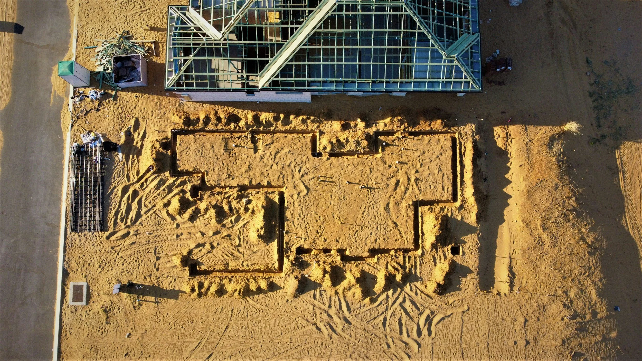 An aerial view of a construction site showing the foundation of a building, surrounding sandy terrain, construction tools, and workers.