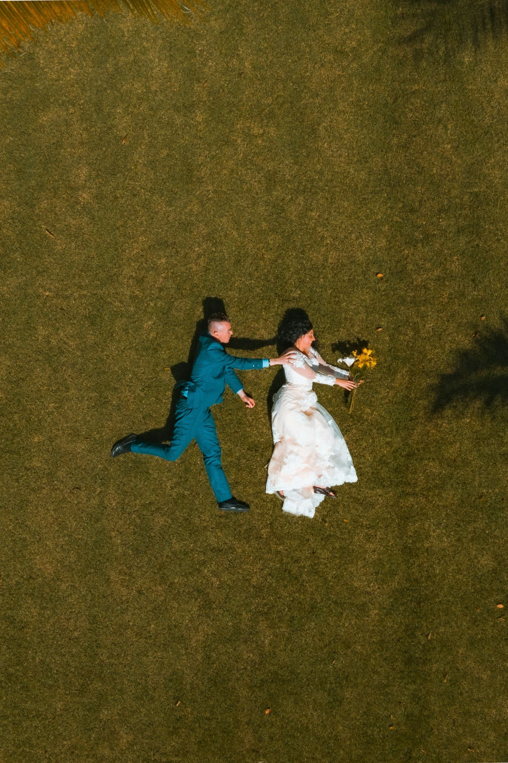 An aerial view of a bride and groom lying on the grass, with the groom reaching out to the bride who is holding a bouquet of flowers.