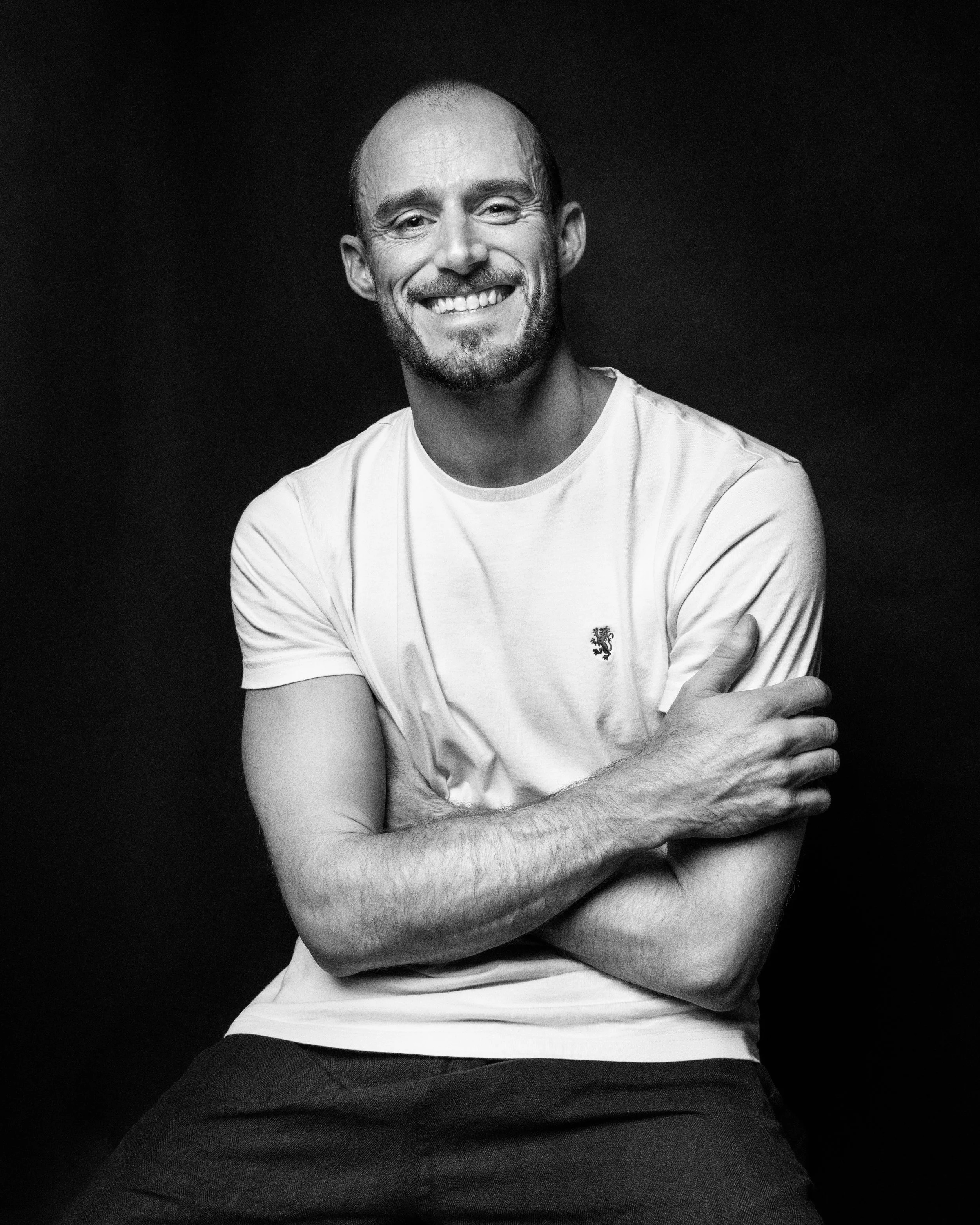 A smiling man with a beard and short hair sitting against a black background, wearing a white T-shirt with a small logo on the chest and dark pants.